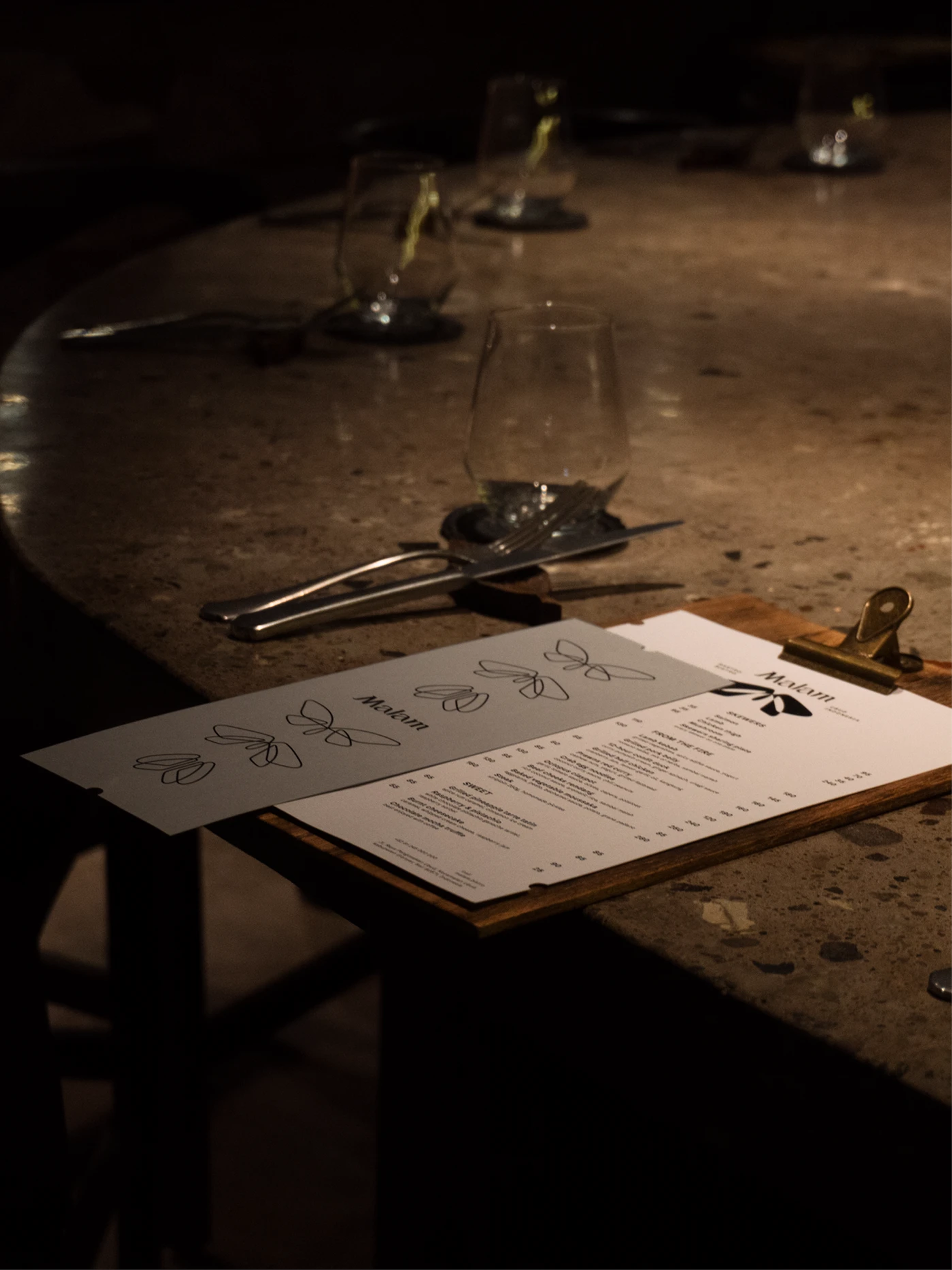 White and grey Malam menus on a brass-clipped wooden board, beside glasses and cutlery on a brown stone counter.