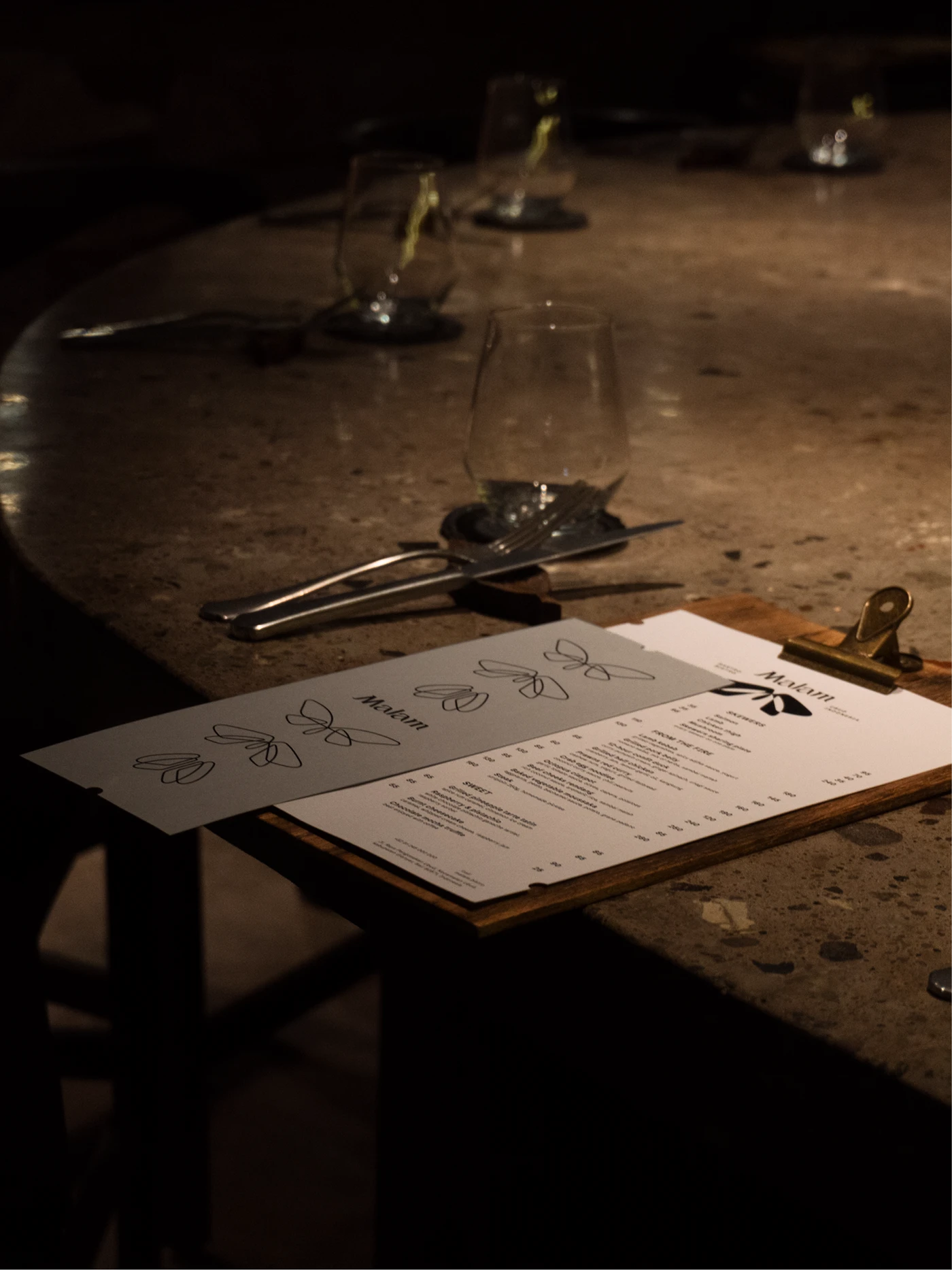 White and grey Malam menus on a brass-clipped wooden board, beside glasses and cutlery on a brown stone counter.