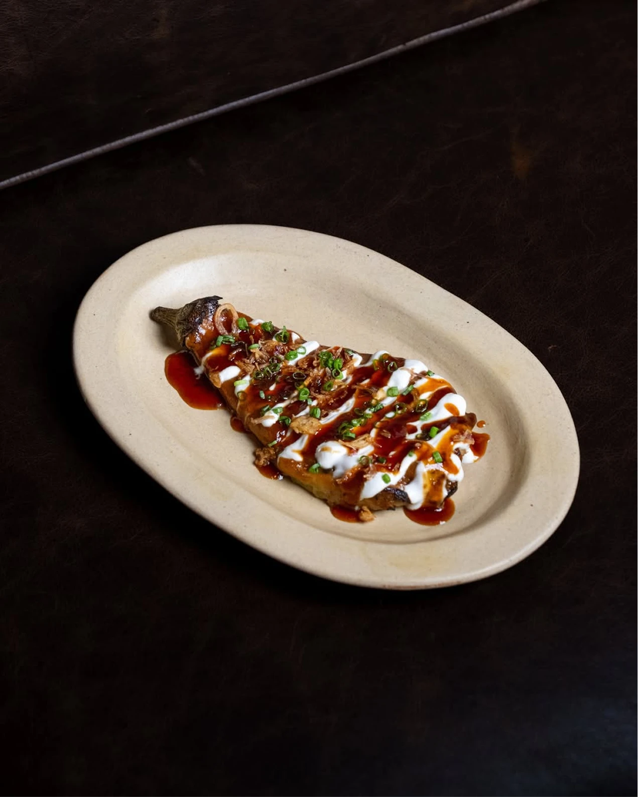 Roasted eggplant topped with brown and white sauces and green onions on a cream oval plate against a dark tabletop.