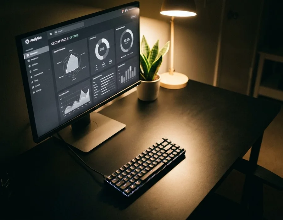 A developer workspace at night with a monitor showing a monochrome dashboard, a mechanical keyboard, a small plant, warm desk-lamp pooling light over black surfaces
