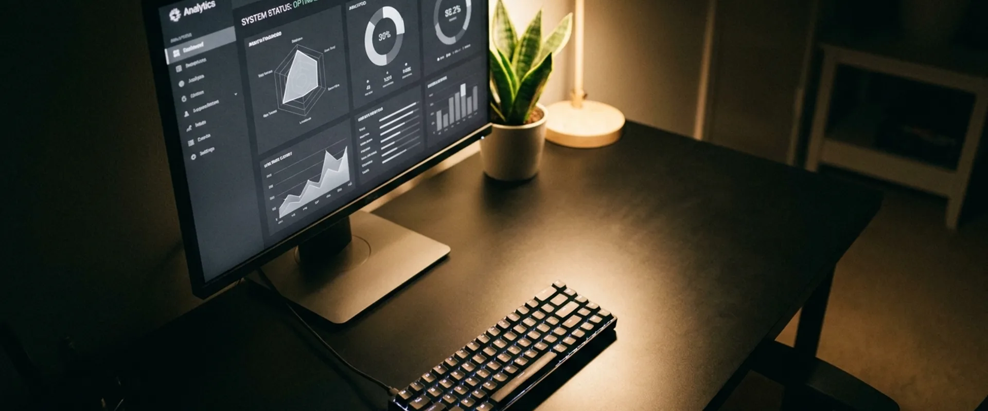 A developer workspace at night with a monitor showing a monochrome dashboard, a mechanical keyboard, a small plant, warm desk-lamp pooling light over black surfaces