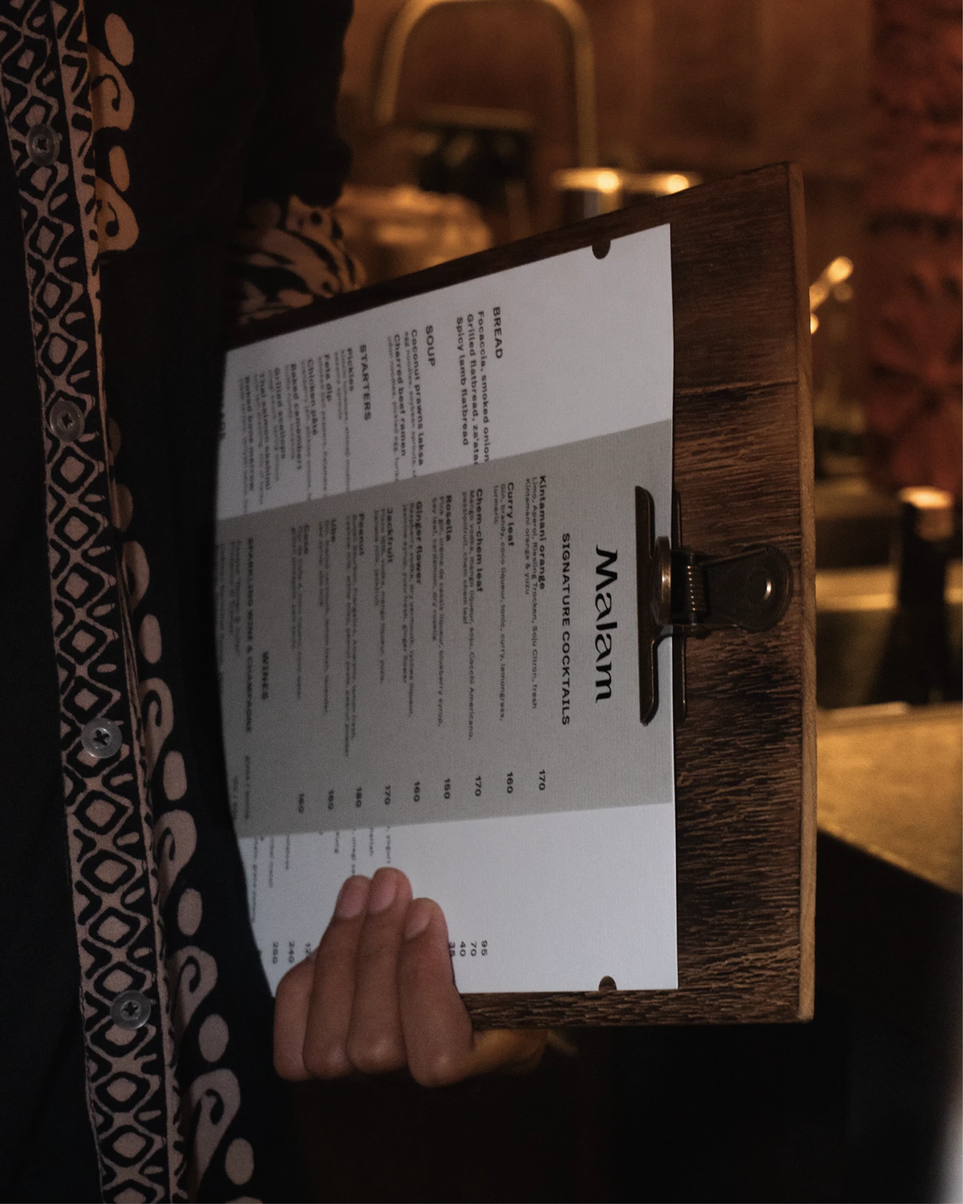 Server holds a wood clipboard with a white and gray “Malam SIGNATURE COCKTAILS” menu in a warm, dim interior.