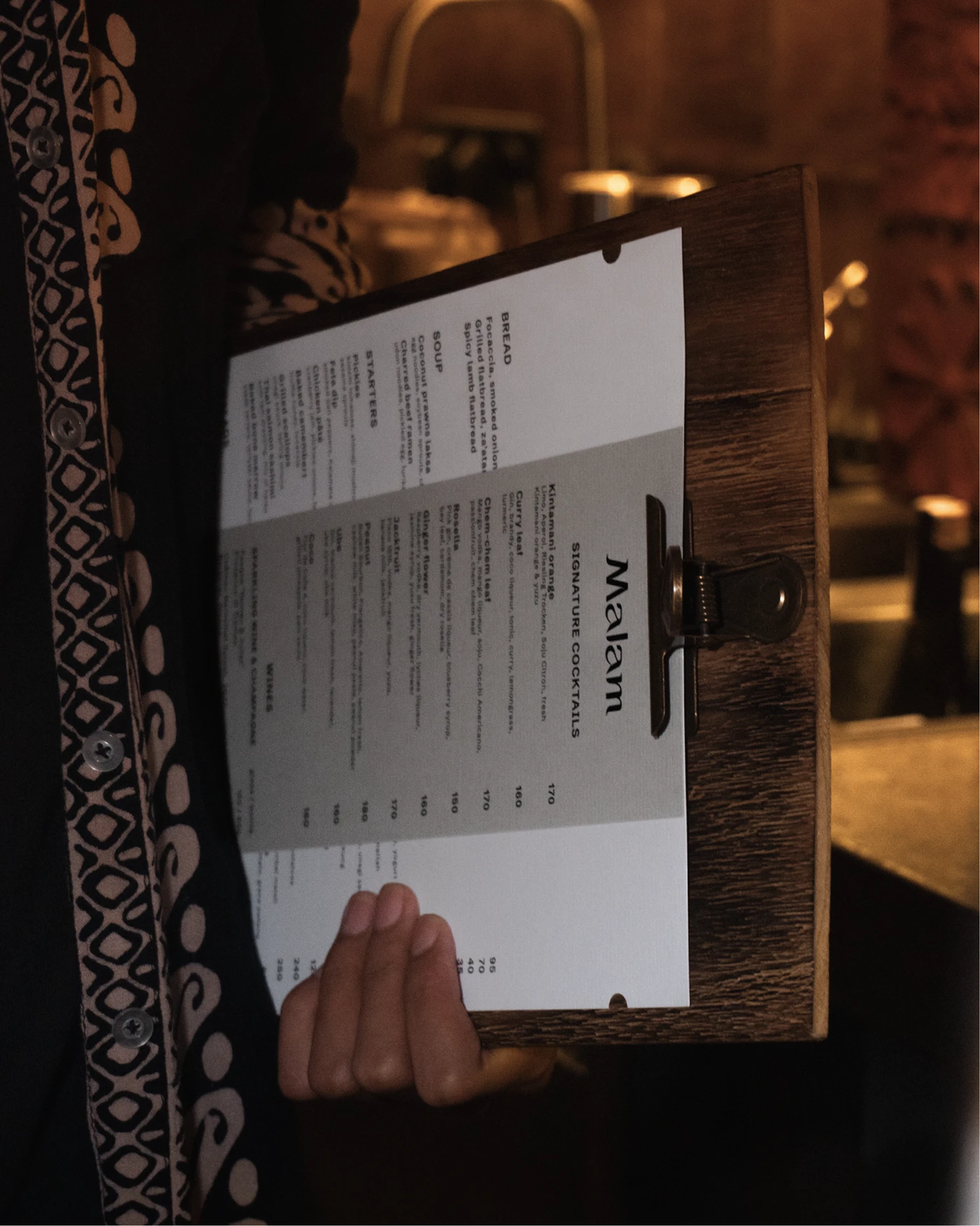 Server holds a wood clipboard with a white and gray “Malam SIGNATURE COCKTAILS” menu in a warm, dim interior.