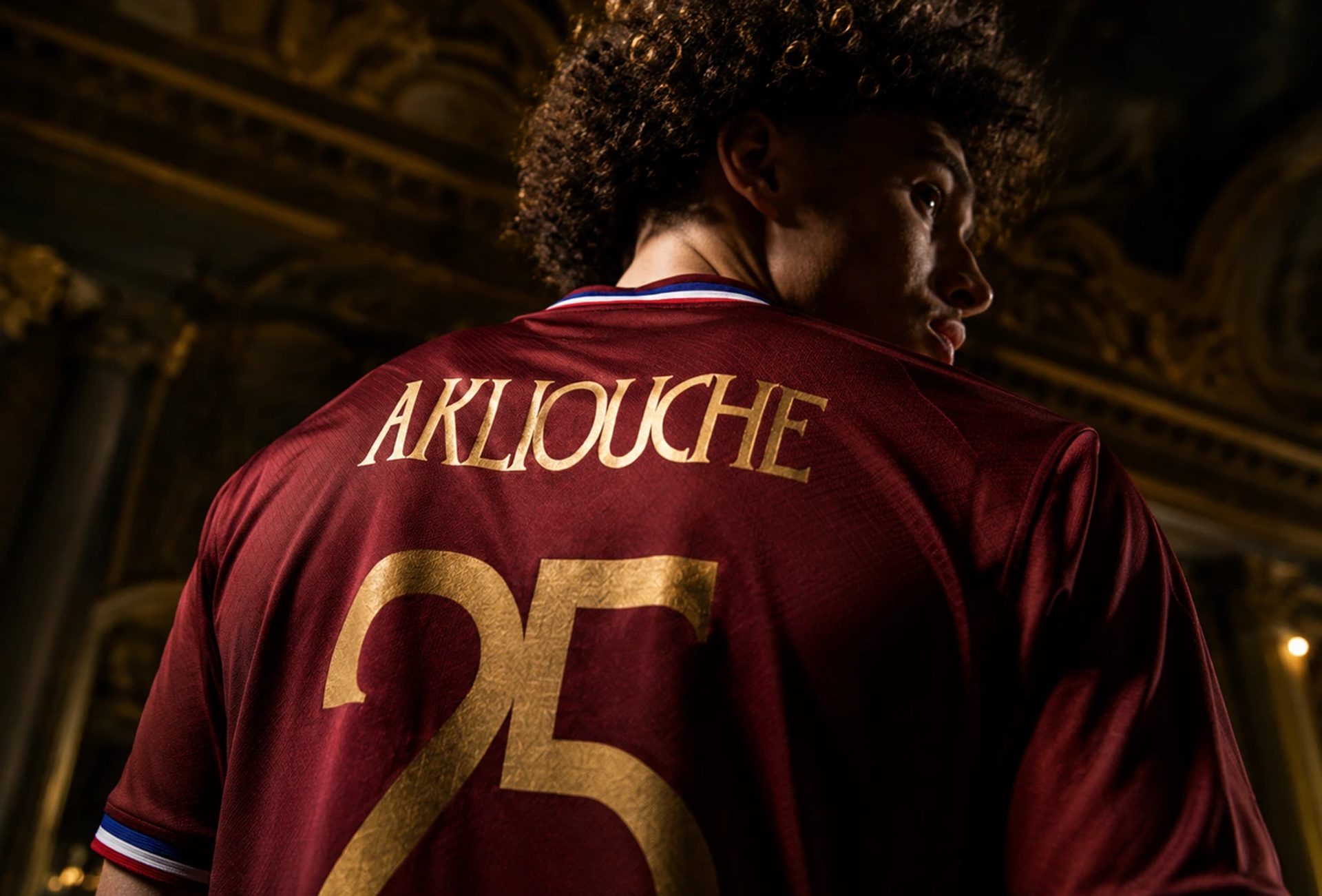 Footballer seen from behind in a burgundy jersey with gold “AKLIOUCHE” and “25”, framed against an ornate dark interior.