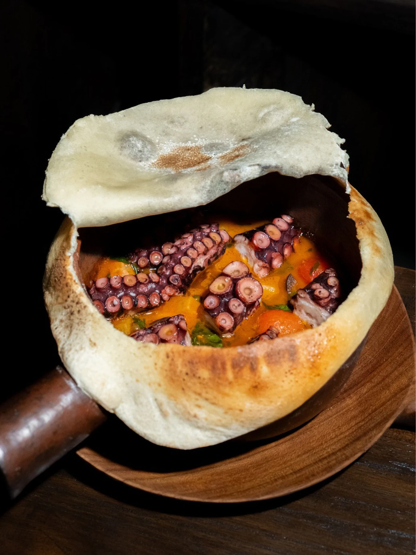 Purple octopus pieces in orange sauce fill a round bread bowl with its lid lifted, served on a wooden plate.