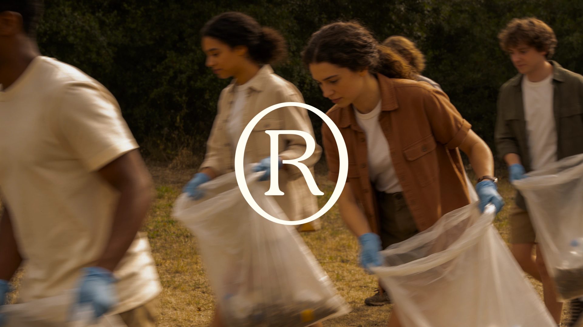 Volunteers in blue gloves hold clear rubbish bags outdoors, with a white circled R logo centred over the scene.