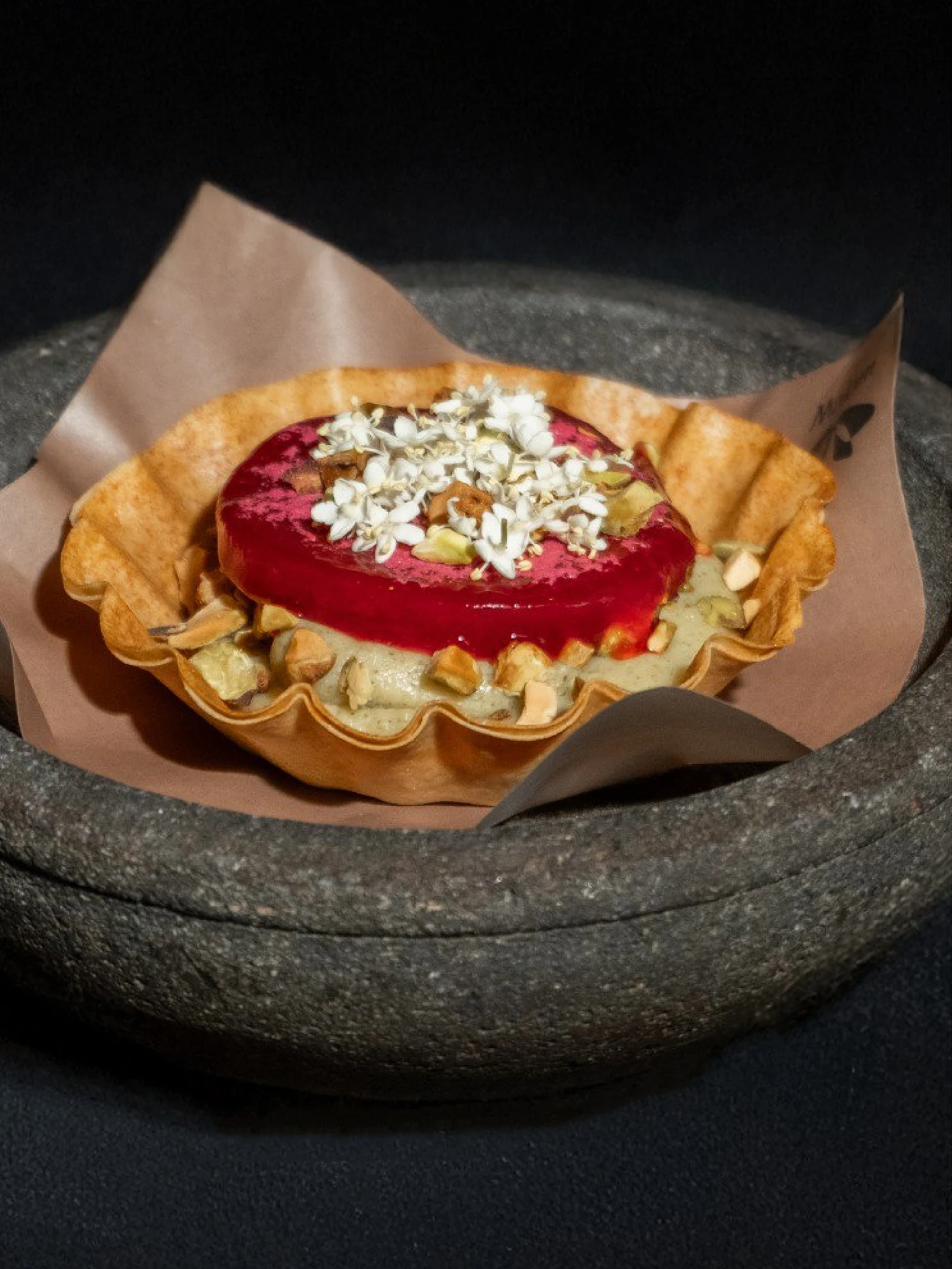Golden pastry bowl with green cream, red jelly, nuts and white petals, served on brown paper in a dark stone dish.