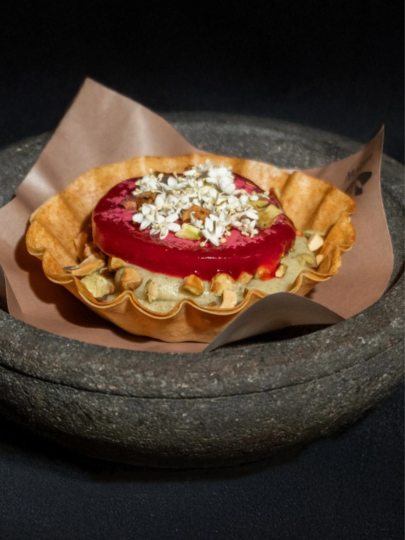 Golden pastry bowl with green cream, red jelly, nuts and white petals, served on brown paper in a dark stone dish.