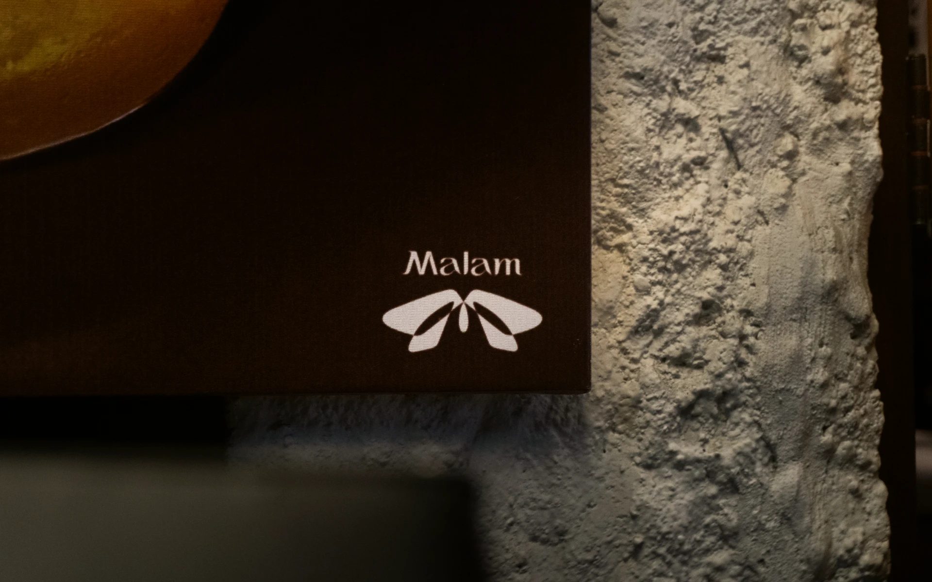Dark brown Malam signage with a white moth symbol mounted beside a rough grey stone wall.