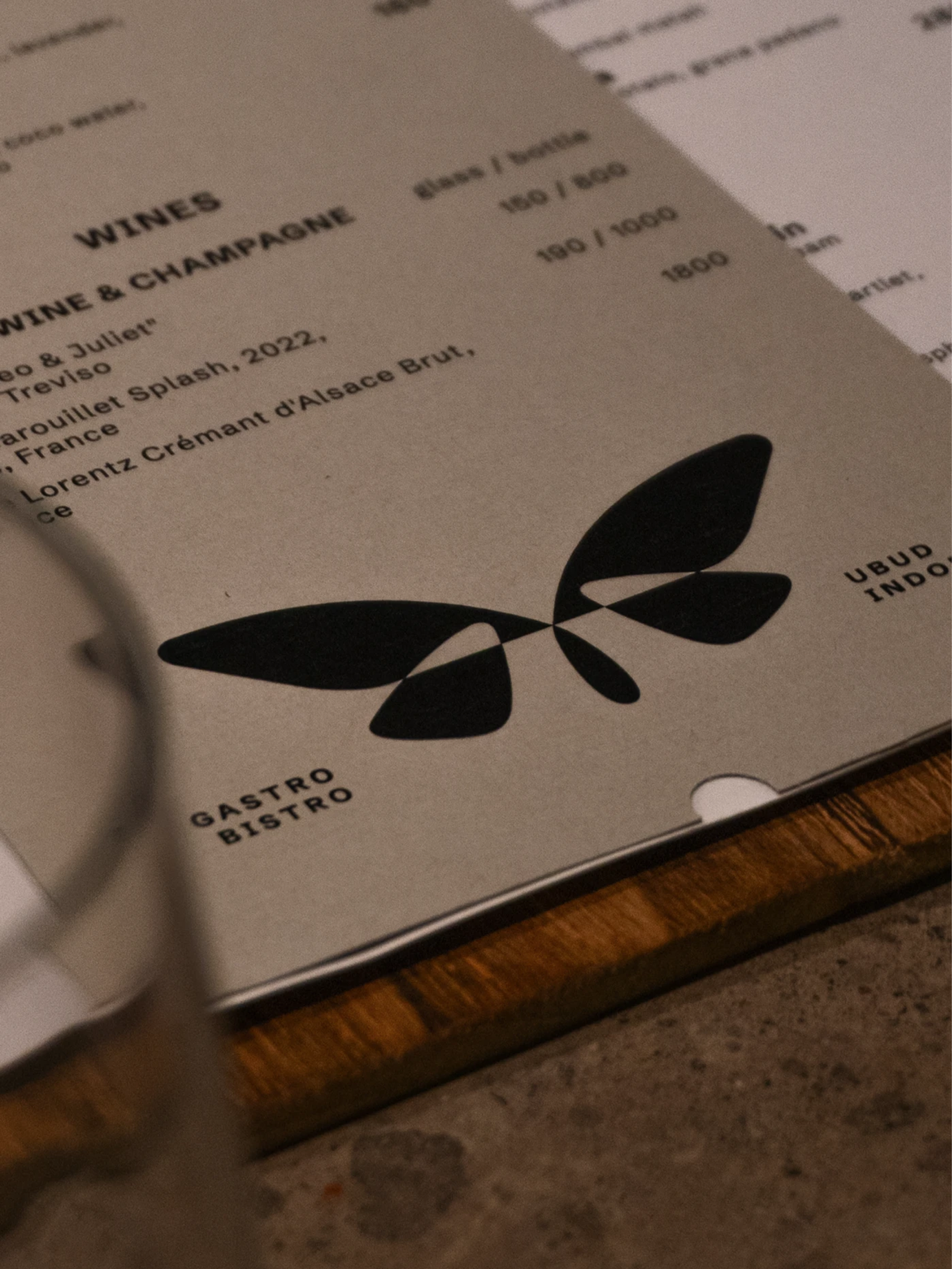 Beige wine menu with black abstract leaf graphics, prices, and “GASTRO BISTRO” on a wooden board beside a blurred glass.