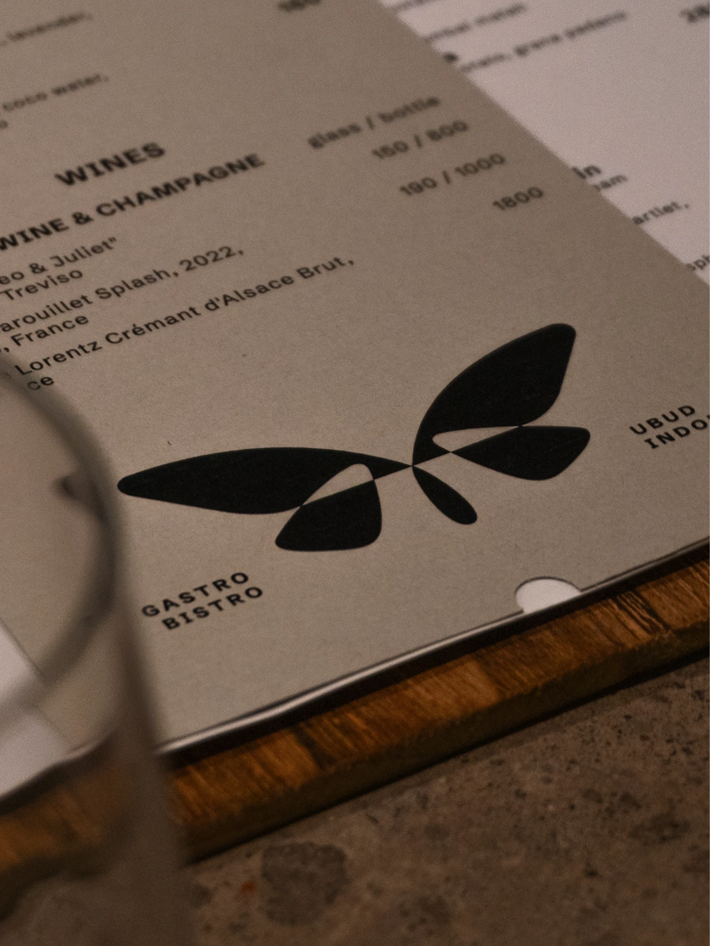 Beige wine menu with black abstract leaf graphics, prices, and “GASTRO BISTRO” on a wooden board beside a blurred glass.
