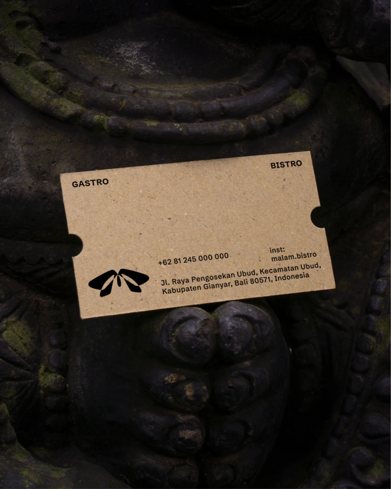 Kraft business card with black “GASTRO” and “BISTRO” text rests diagonally on a dark moss-covered stone carving.
