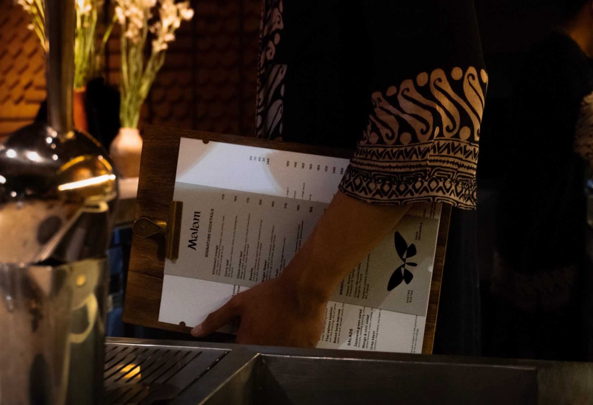 Server holds a wooden clipboard with a black-and-white Malam menu beside a metal ice bucket in warm amber light.