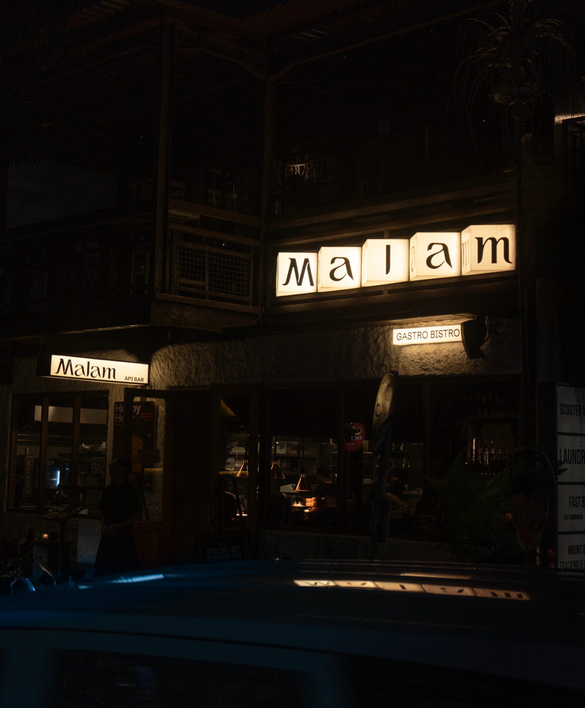 Cream illuminated signs reading “Malam,” “API BAR” and “GASTRO BISTRO” mark a dark restaurant facade at night.