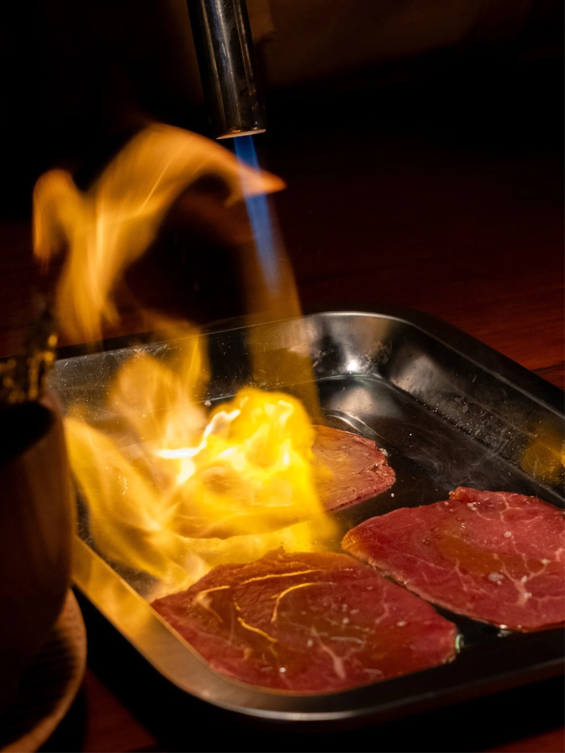 Blue blowtorch flame and orange fire sear slices of red beef in a black metal tray against a dark background.