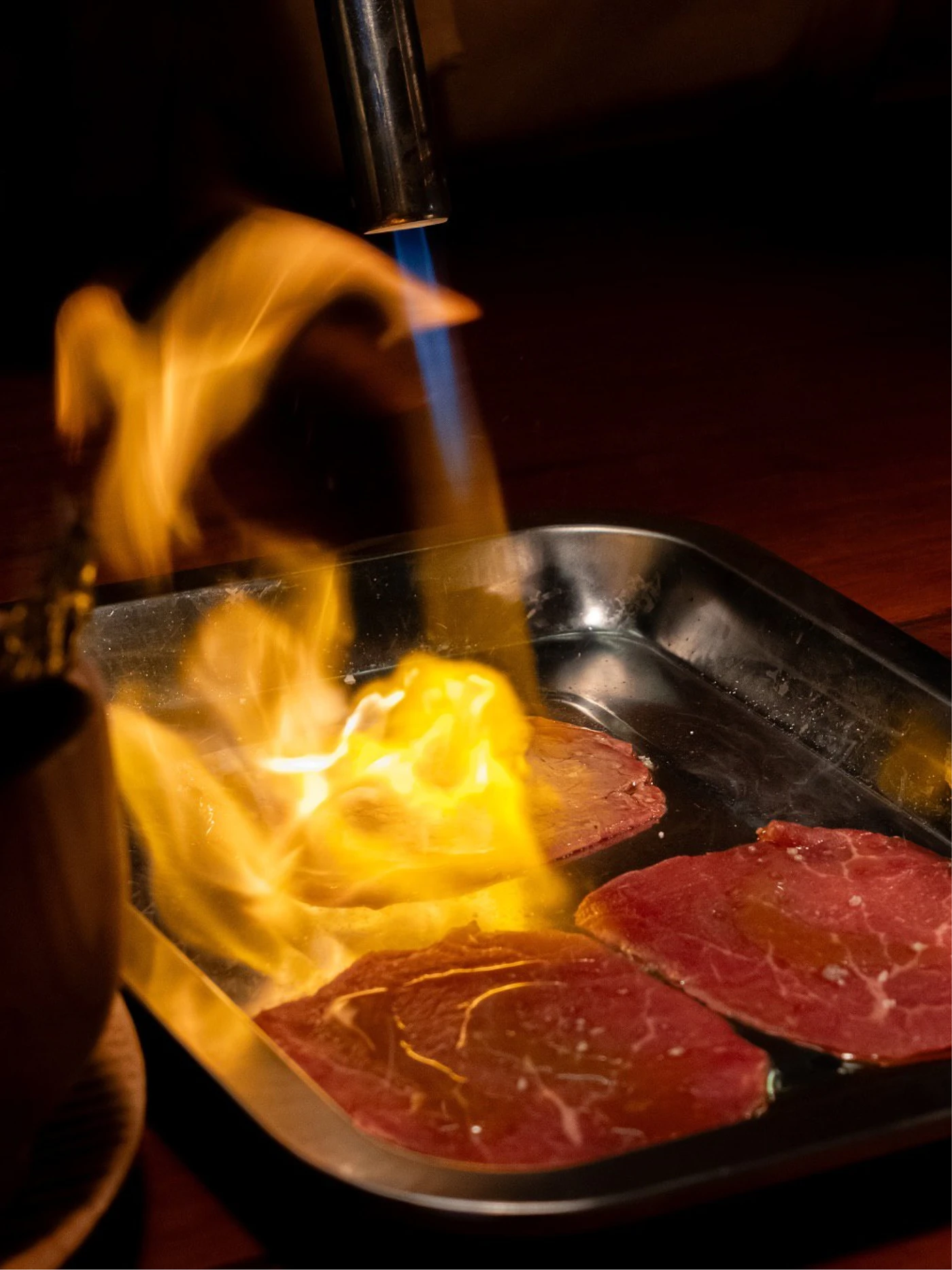 Blue blowtorch flame and orange fire sear slices of red beef in a black metal tray against a dark background.