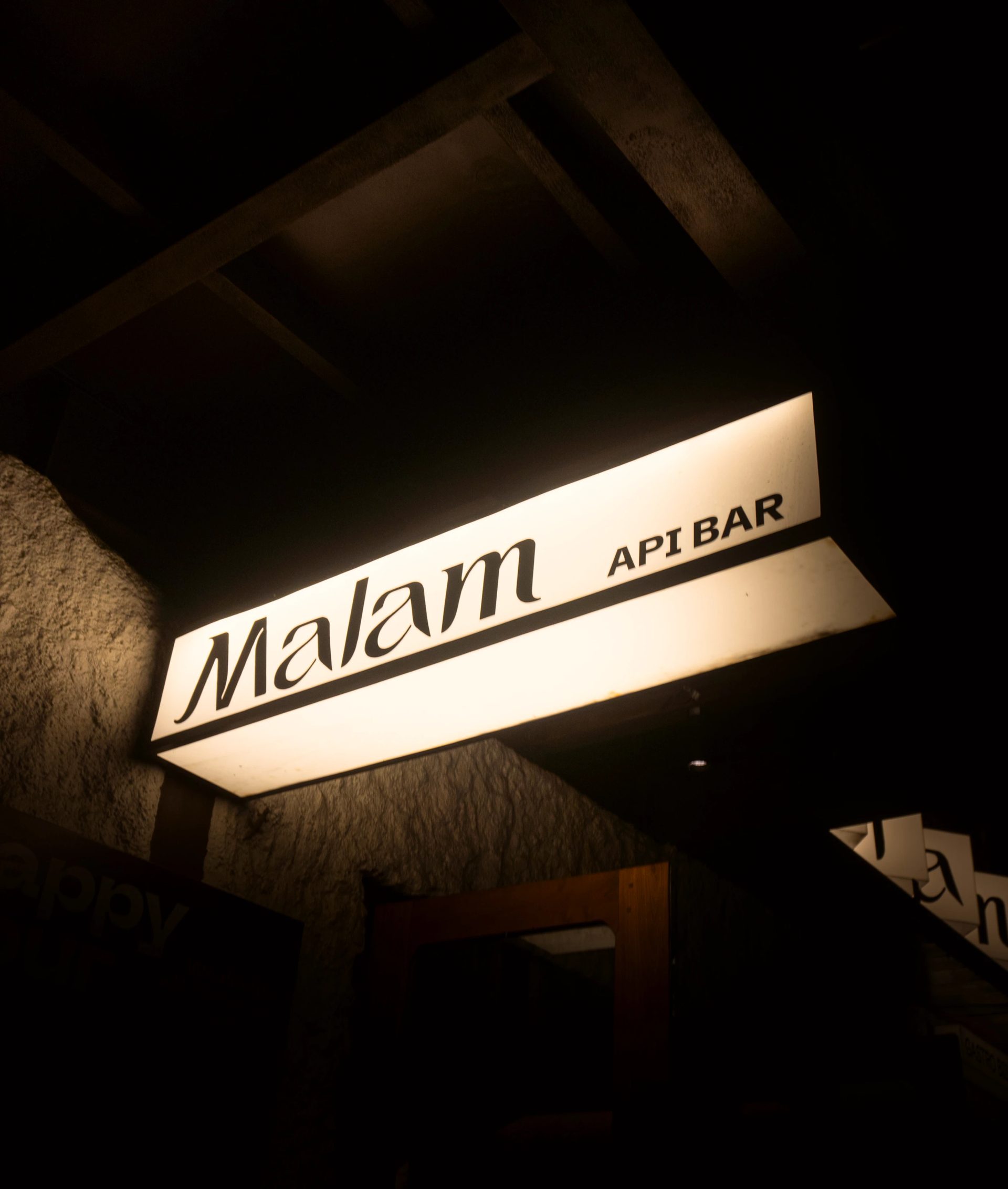 Illuminated cream projecting sign reading “Malam API BAR” in black lettering against a dark exterior at night.