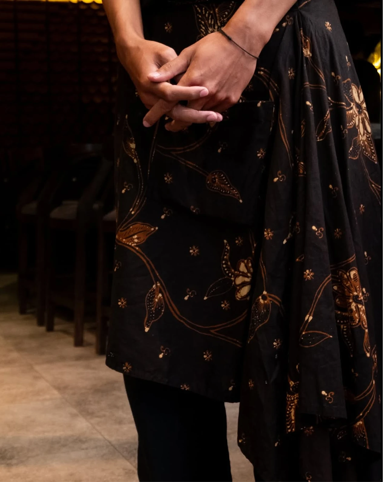 Close-up of clasped hands over a black uniform with brown floral motifs, set against a dim bistro interior.