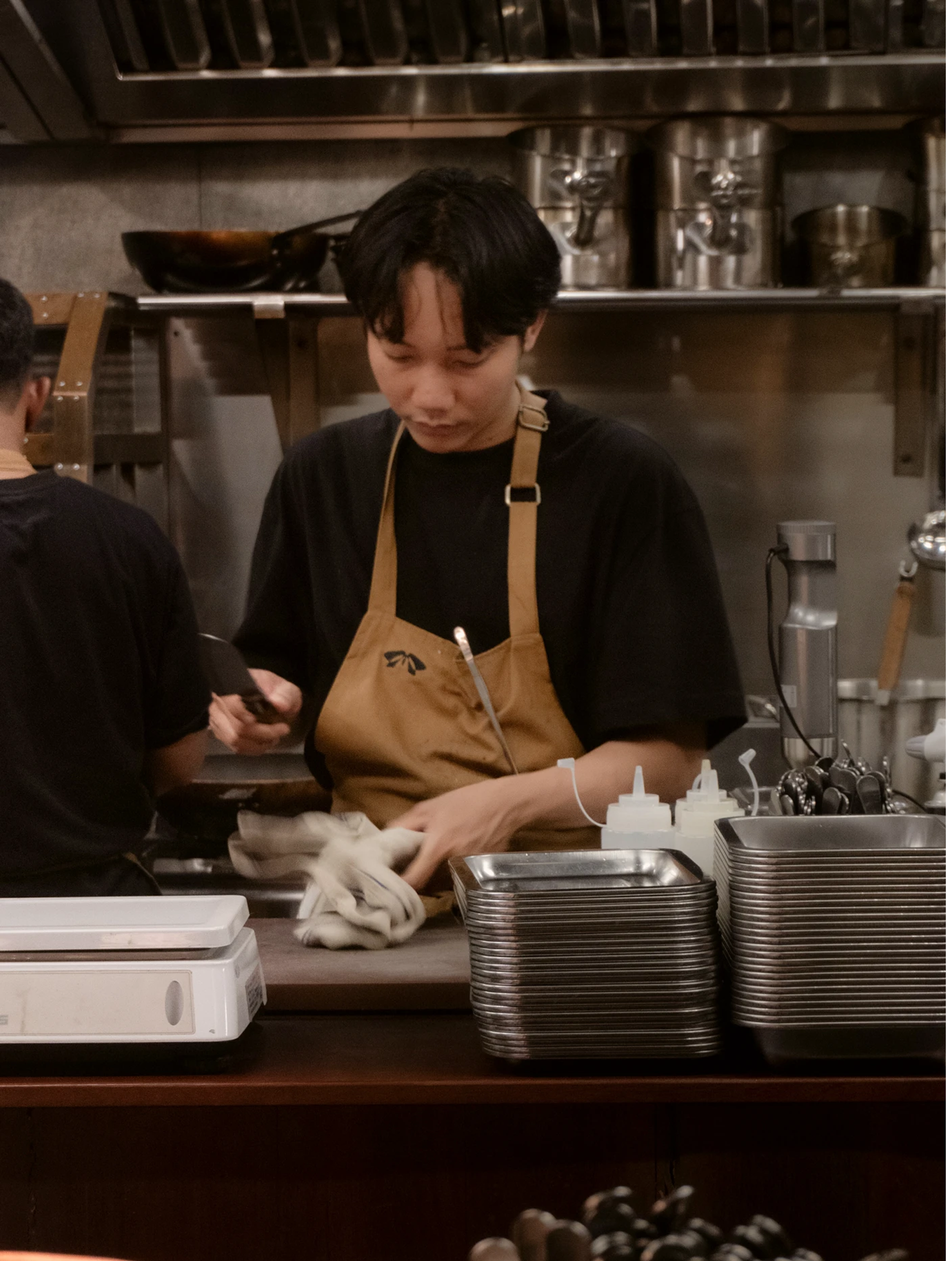 Chef in a tan apron works at a stainless steel bistro counter beside stacked metal trays and kitchen equipment.