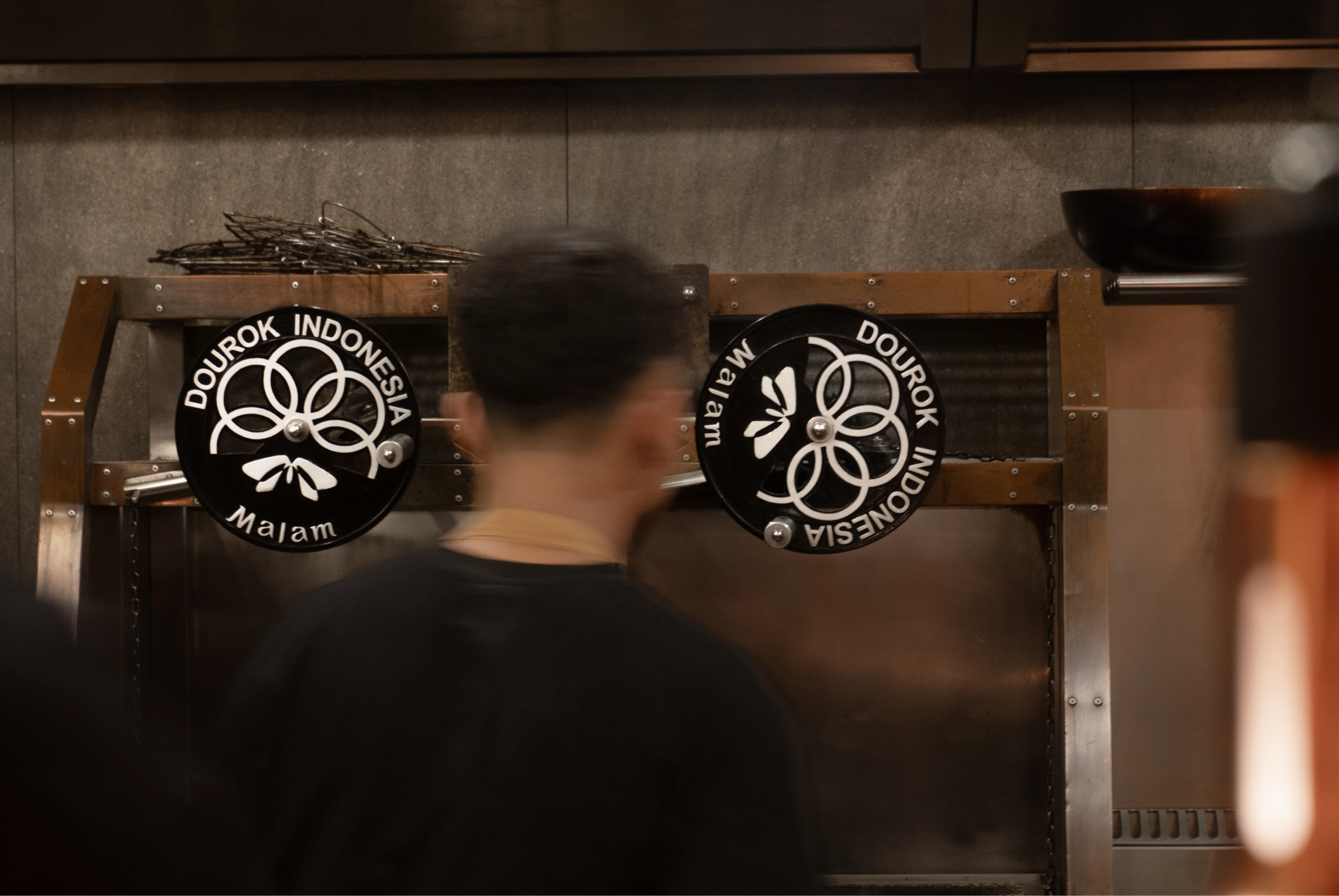Two black circular signs reading “DOUROK INDONESIA” and “Malam” hang on a brown metal kitchen frame behind a blurred cook.