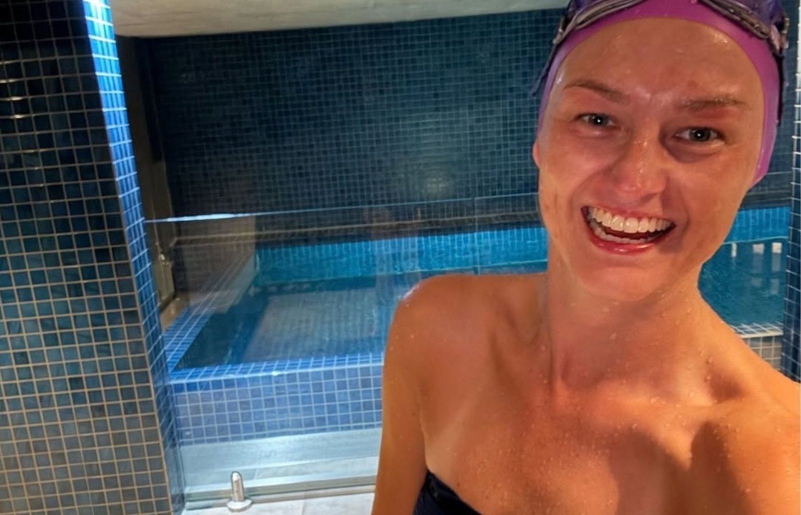 Jen smiling beside an indoor pool wearing a purple swim cap and goggles