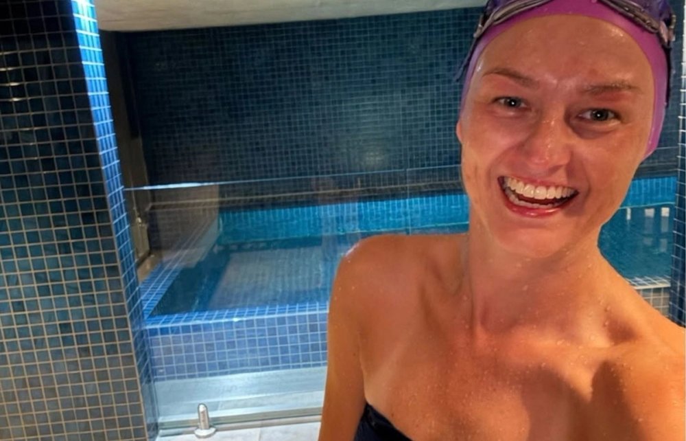 Jen smiling beside an indoor pool wearing a purple swim cap and goggles