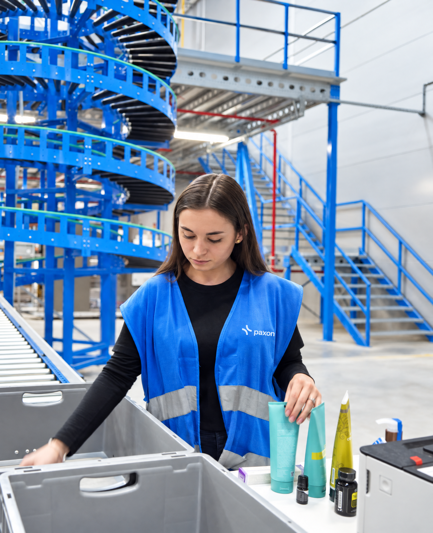 A woman in a blue Paxon vest sorts products into bins in a warehouse with blue spiral conveyors.