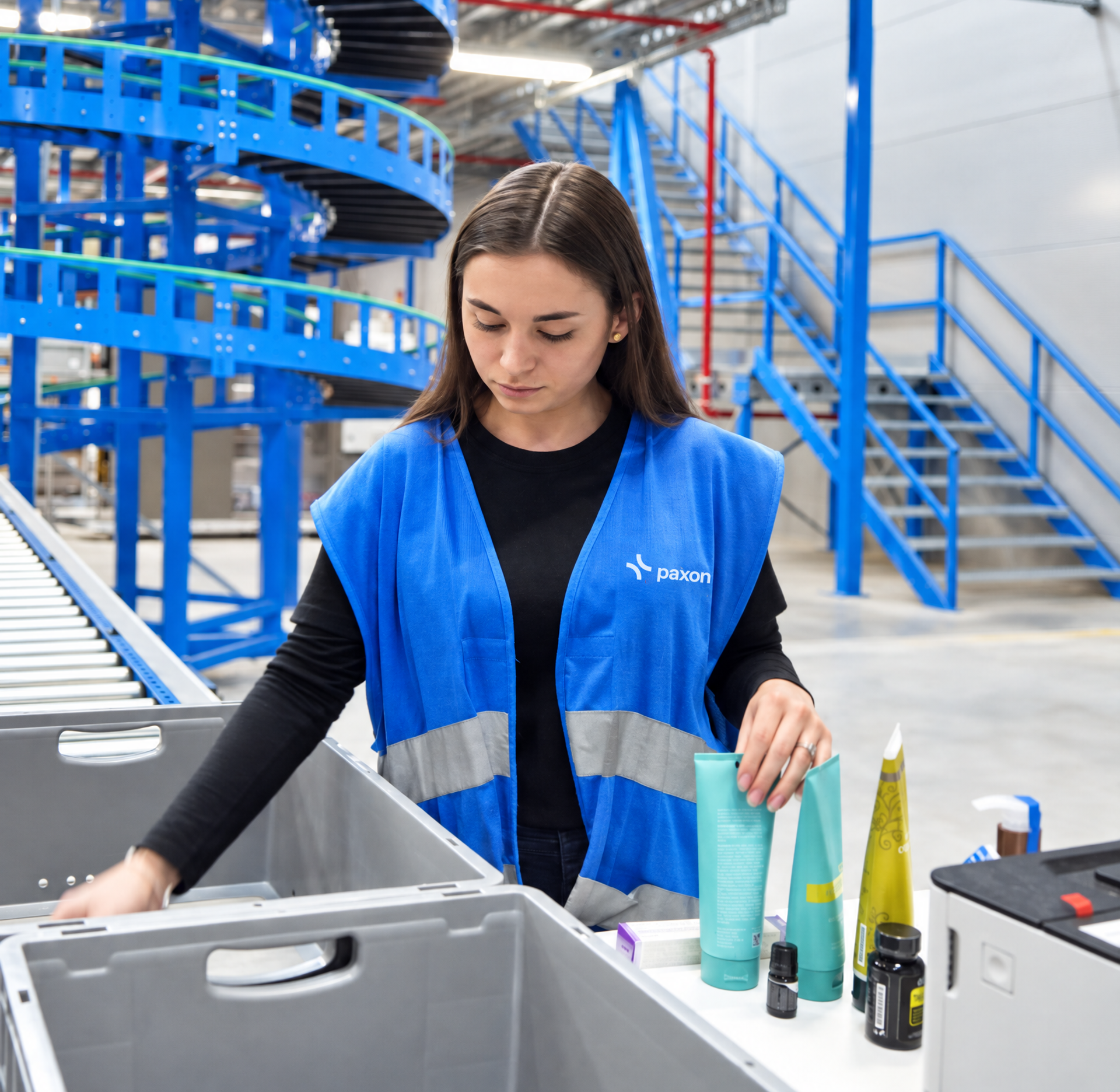 A woman in a blue Paxon vest sorts products into bins in a warehouse with blue spiral conveyors.