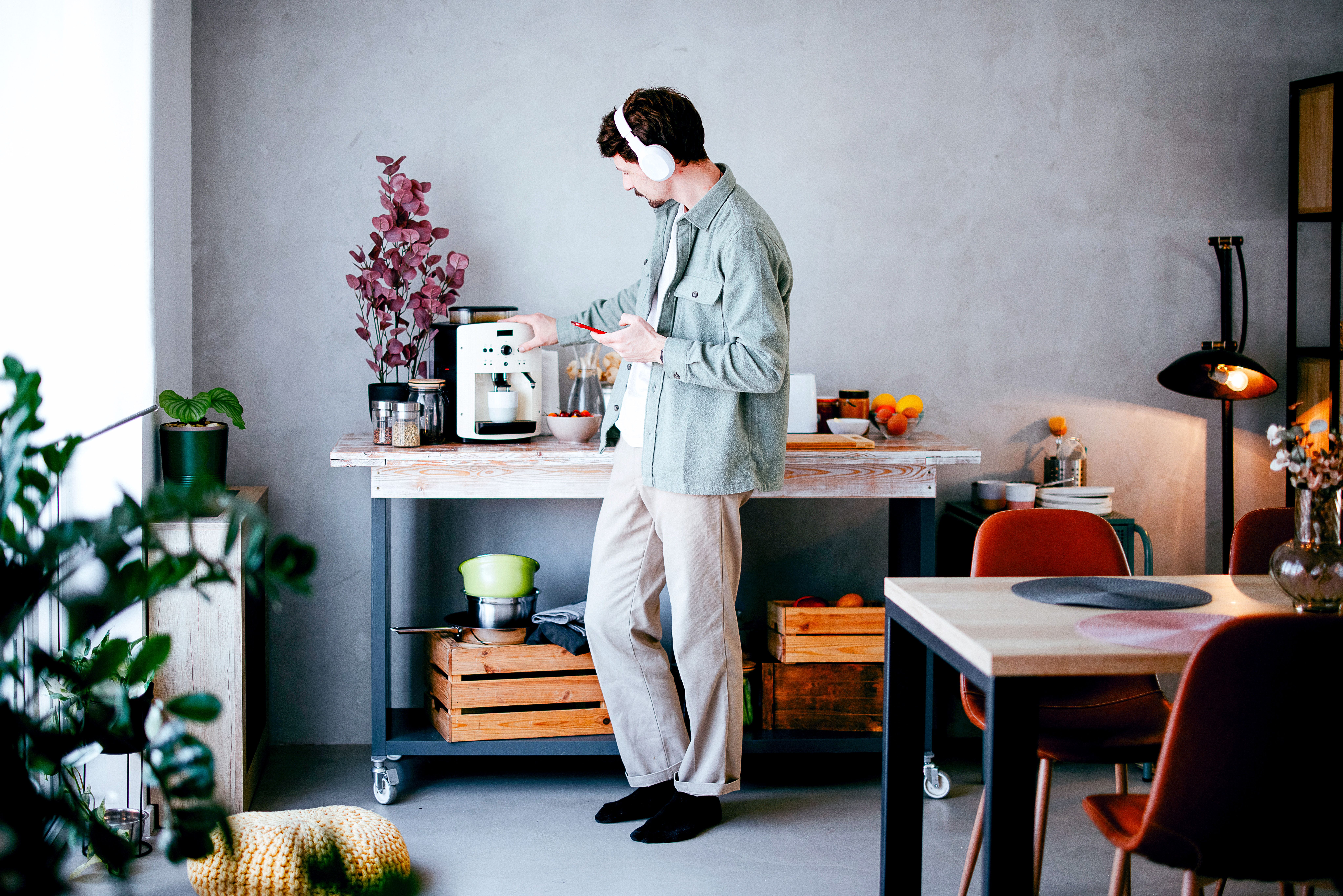 A man wearing headphones and using a smartphone prepares coffee at a kitchen cart.