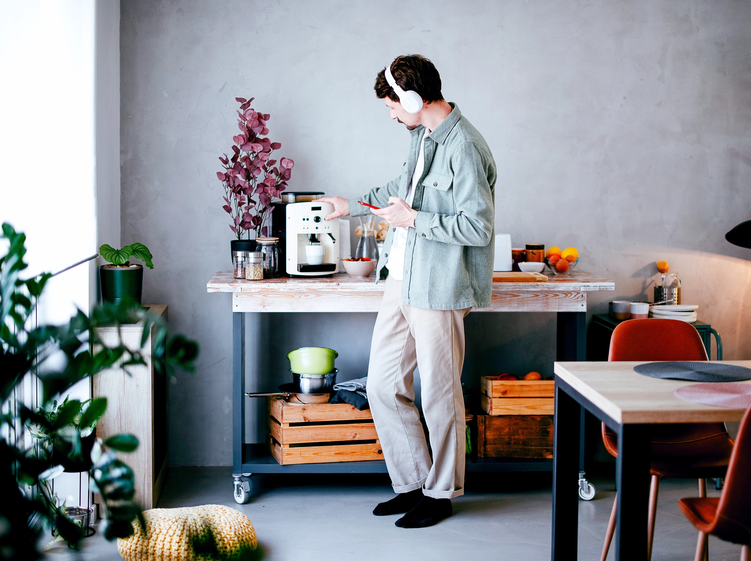A man wearing headphones and using a smartphone prepares coffee at a kitchen cart.