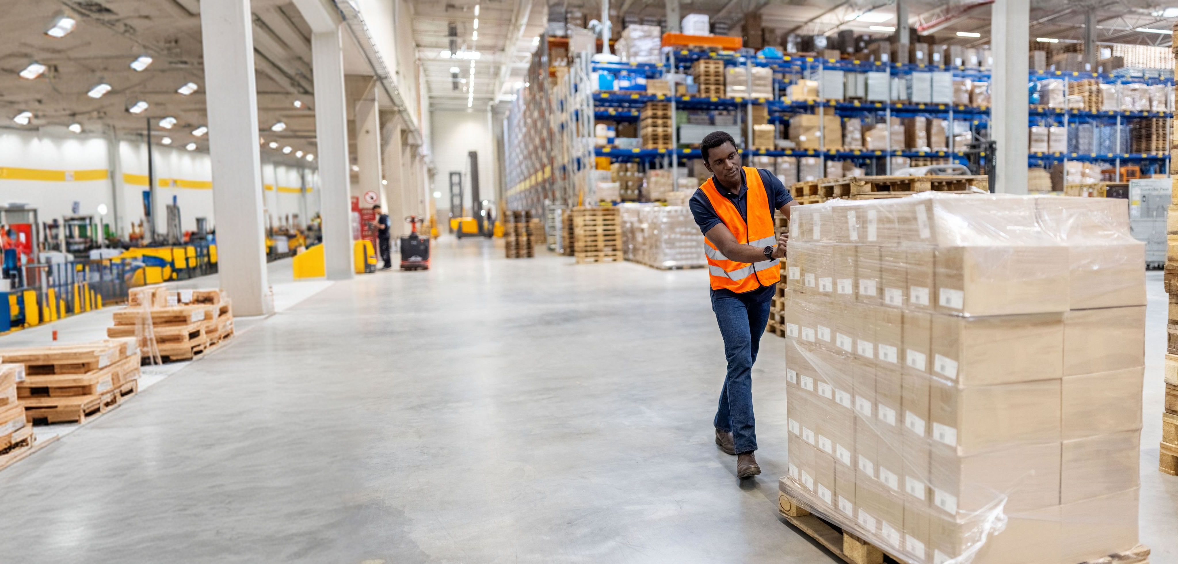 A man in an orange safety vest pushes a pallet of boxes through a large warehouse.