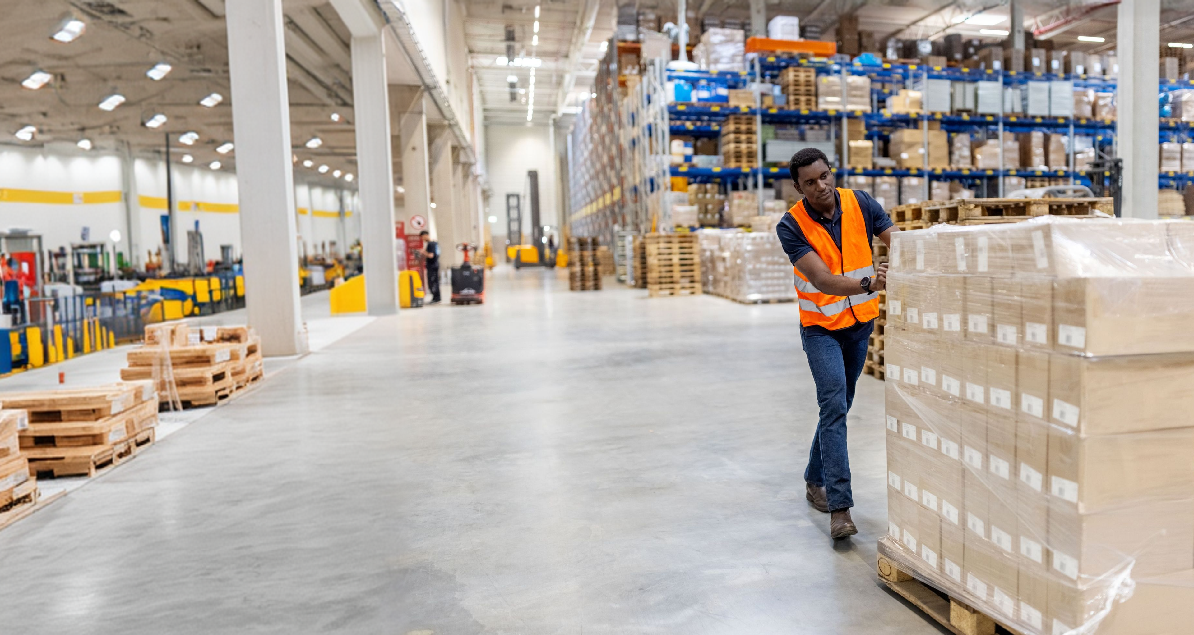 Man in a safety vest pushing boxes on a pallet in a large warehouse.
