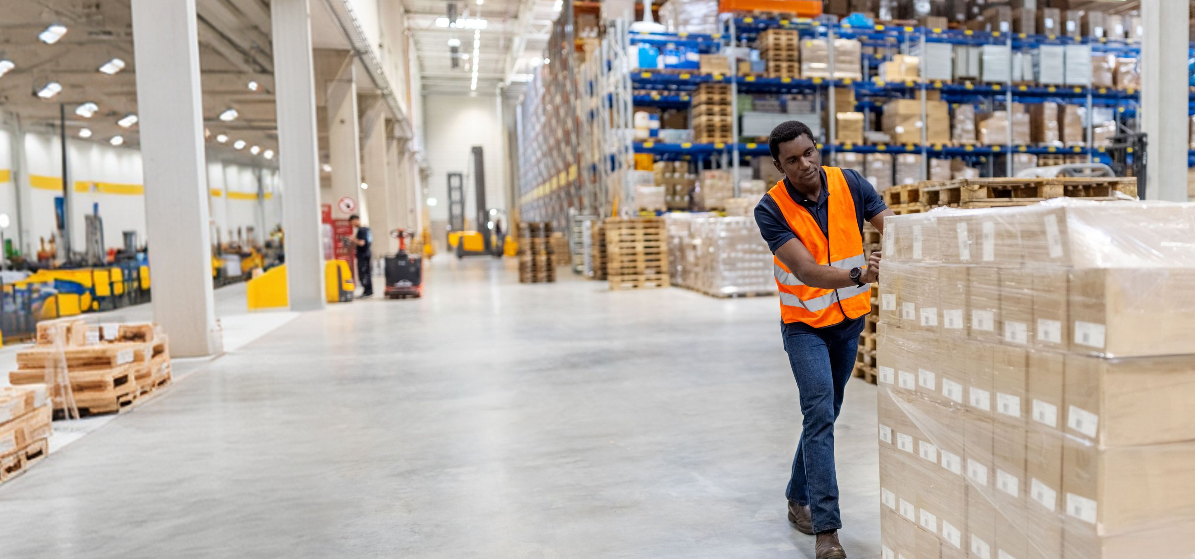 An African American man in an orange safety vest pushes a stack of boxes in a large warehouse.