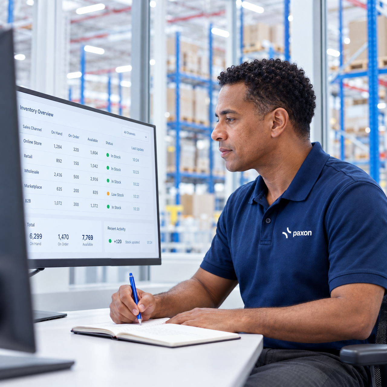 A man in a blue polo shirt monitors inventory on a computer screen and writes in a notebook, with a warehouse behind him.