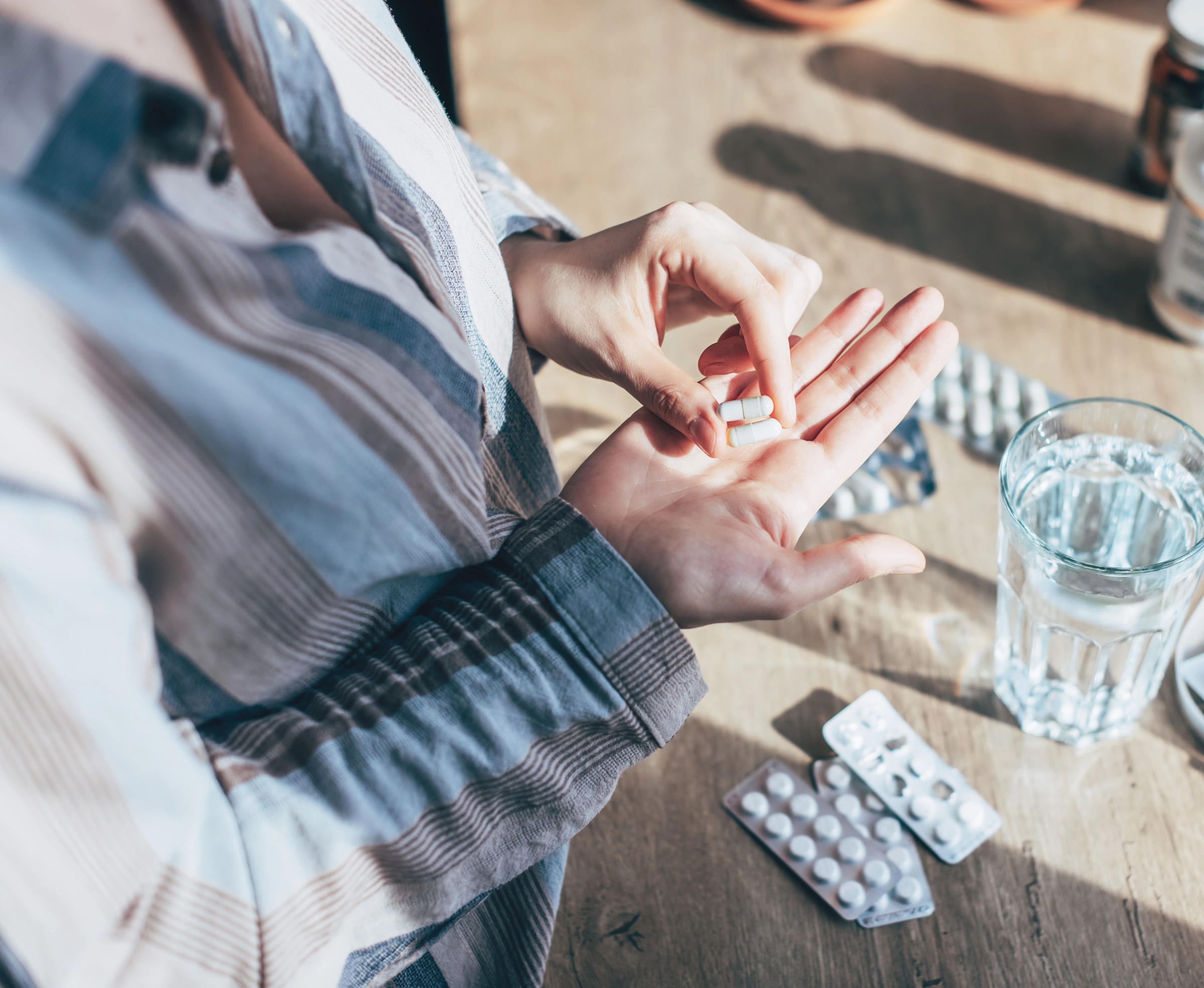 A person's hands holding pills over a wooden table with a glass of water and various medication packs.