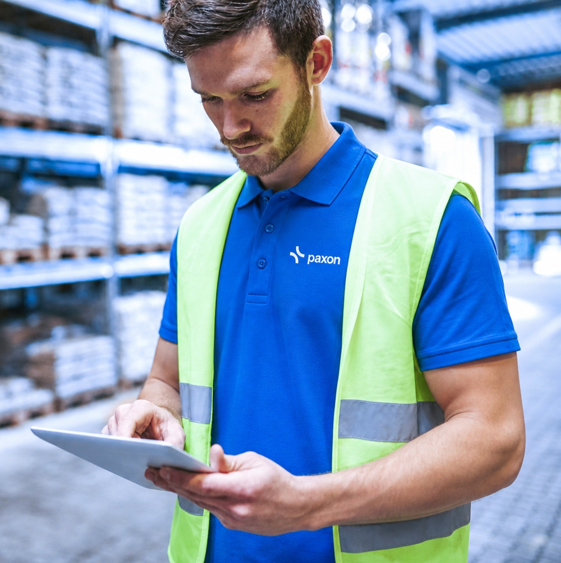 A man in a blue Paxon uniform and a yellow high-vis vest uses a tablet in a warehouse.