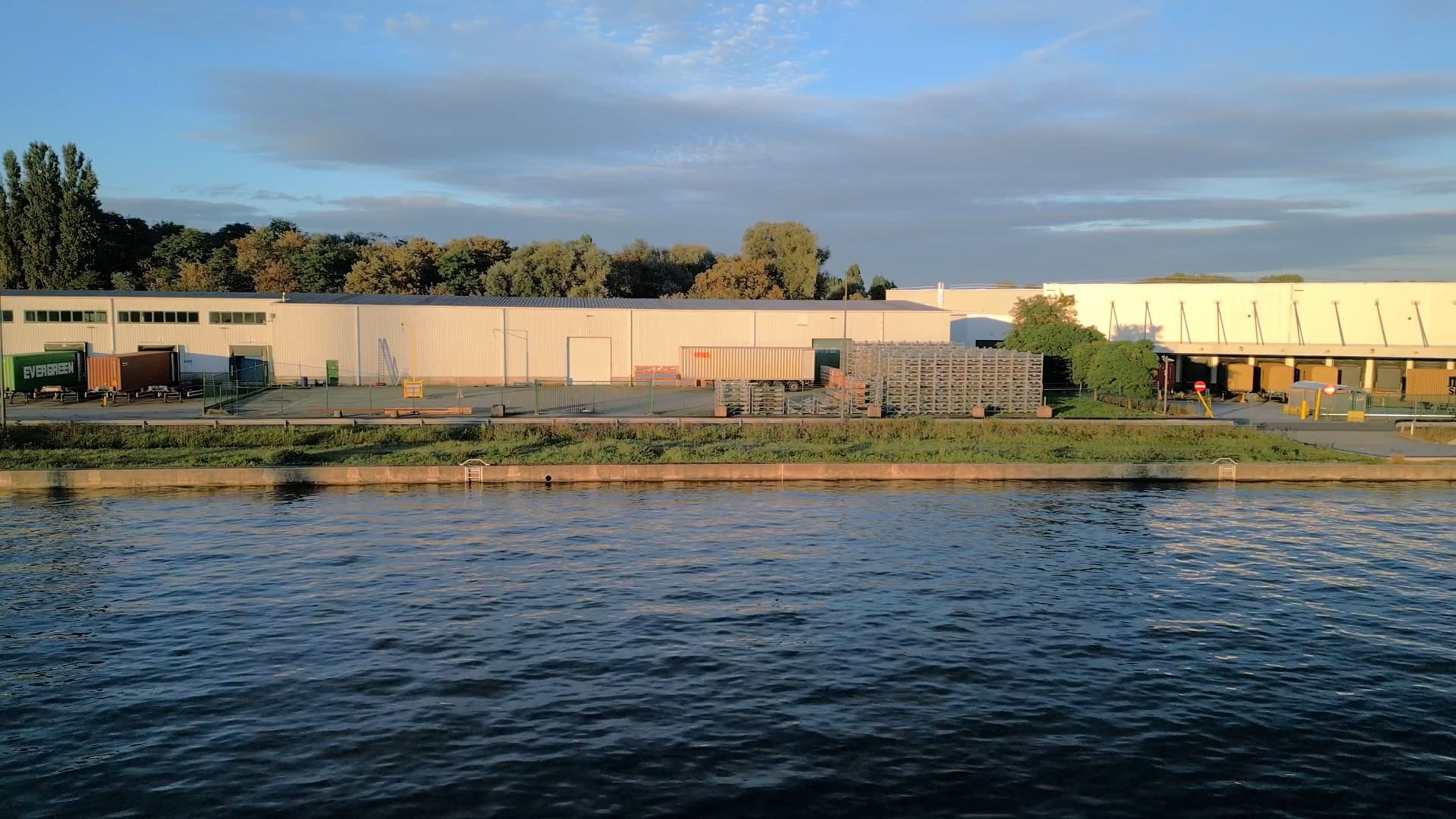 Een industrieel complex aan het water met laadperrons, omringd door bomen, onder een deels bewolkte lucht.