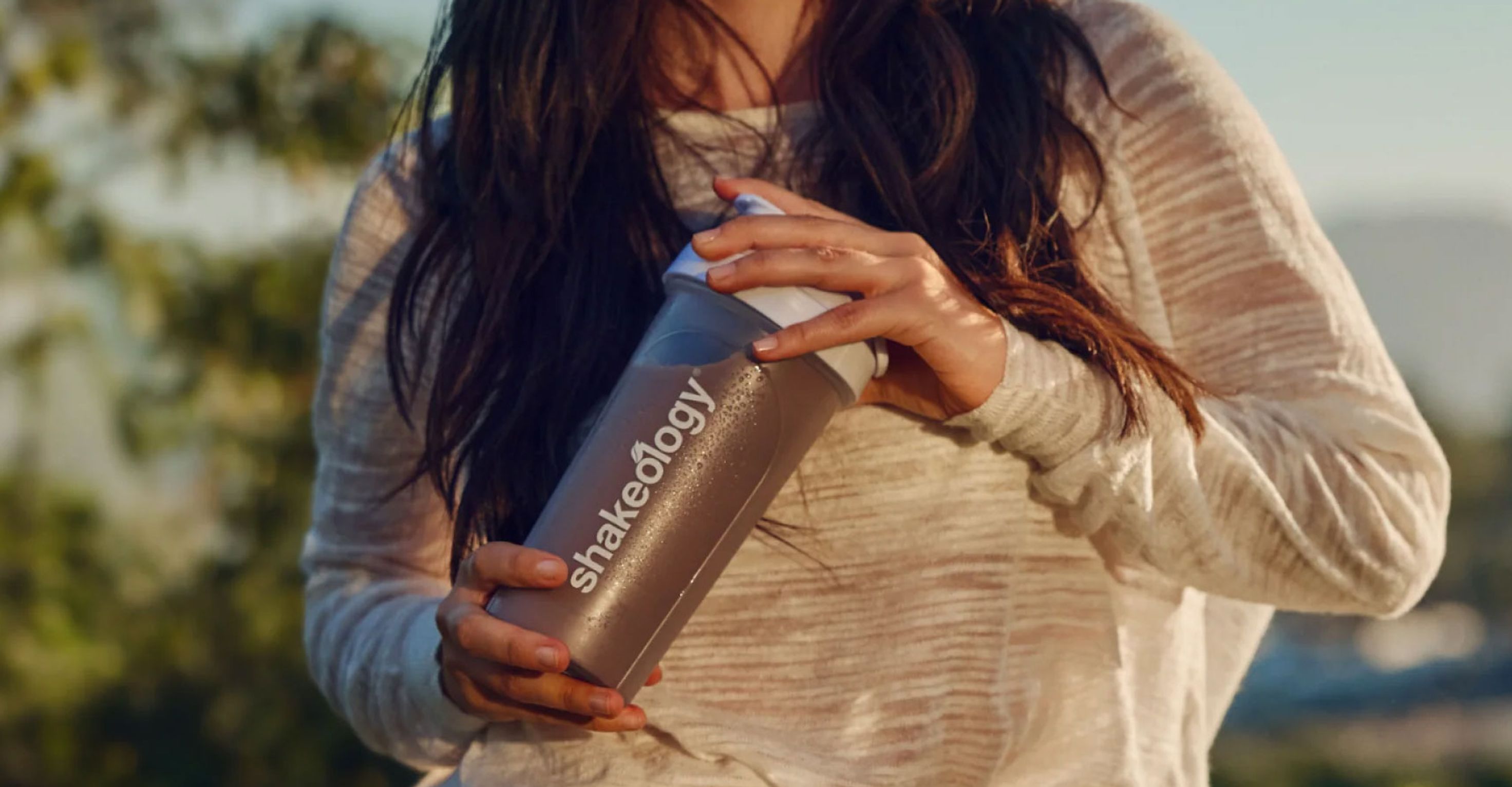 A person holds a Shakeology shaker bottle outdoors.