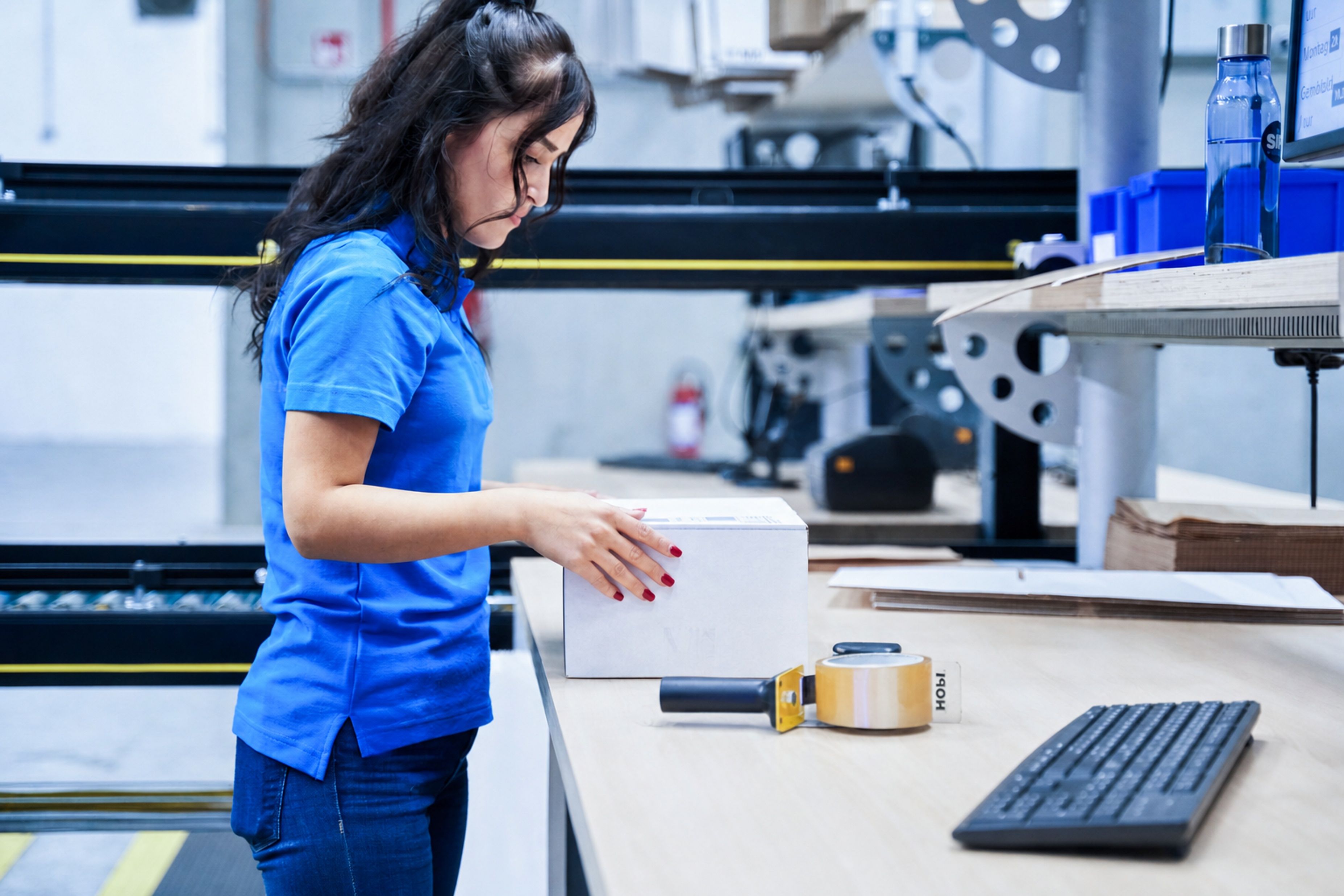 Une femme en polo bleu ferme un colis blanc à un poste de travail.