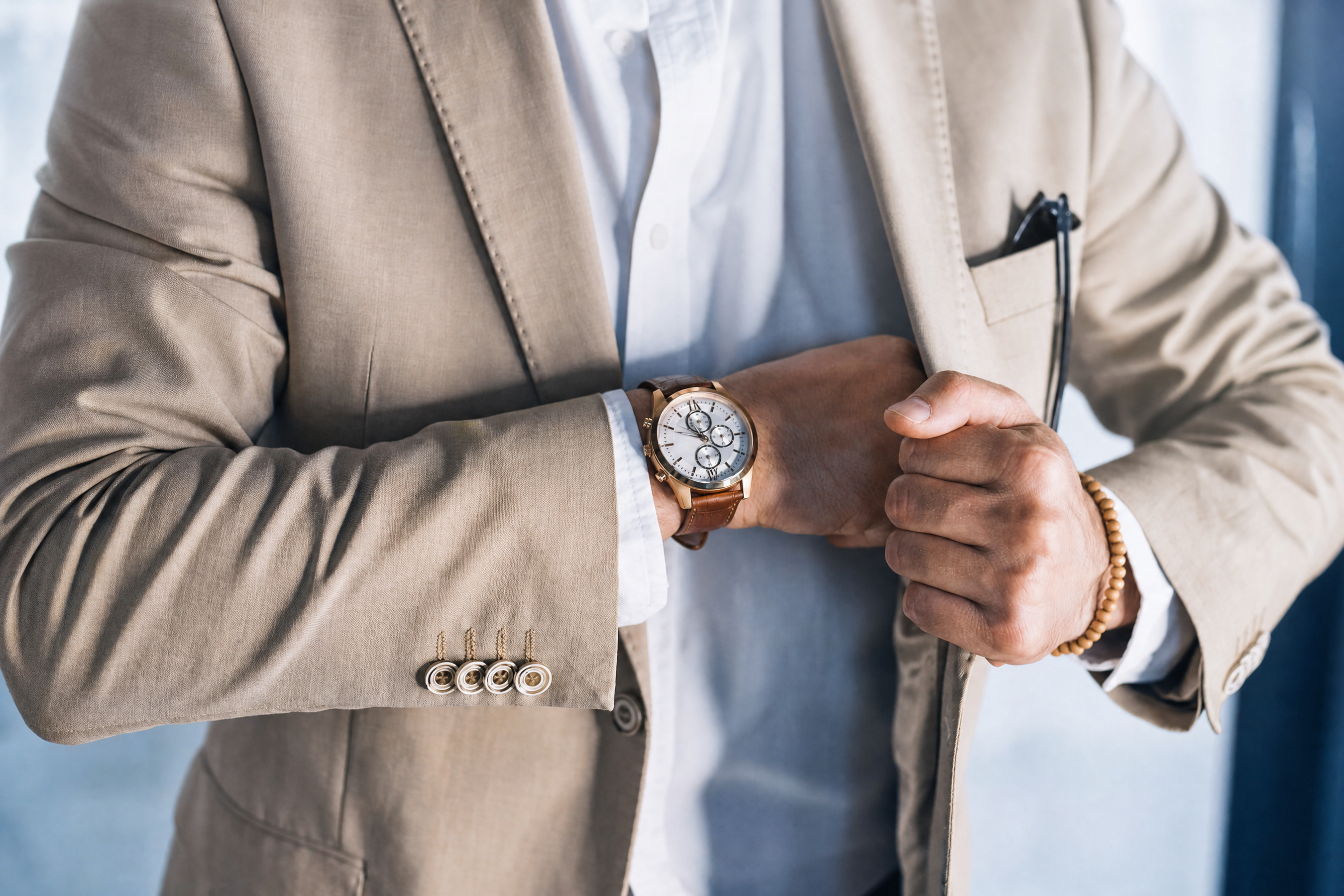Close-up of a person in a beige blazer, white shirt, and wristwatch, with a hand tucked into their jacket.