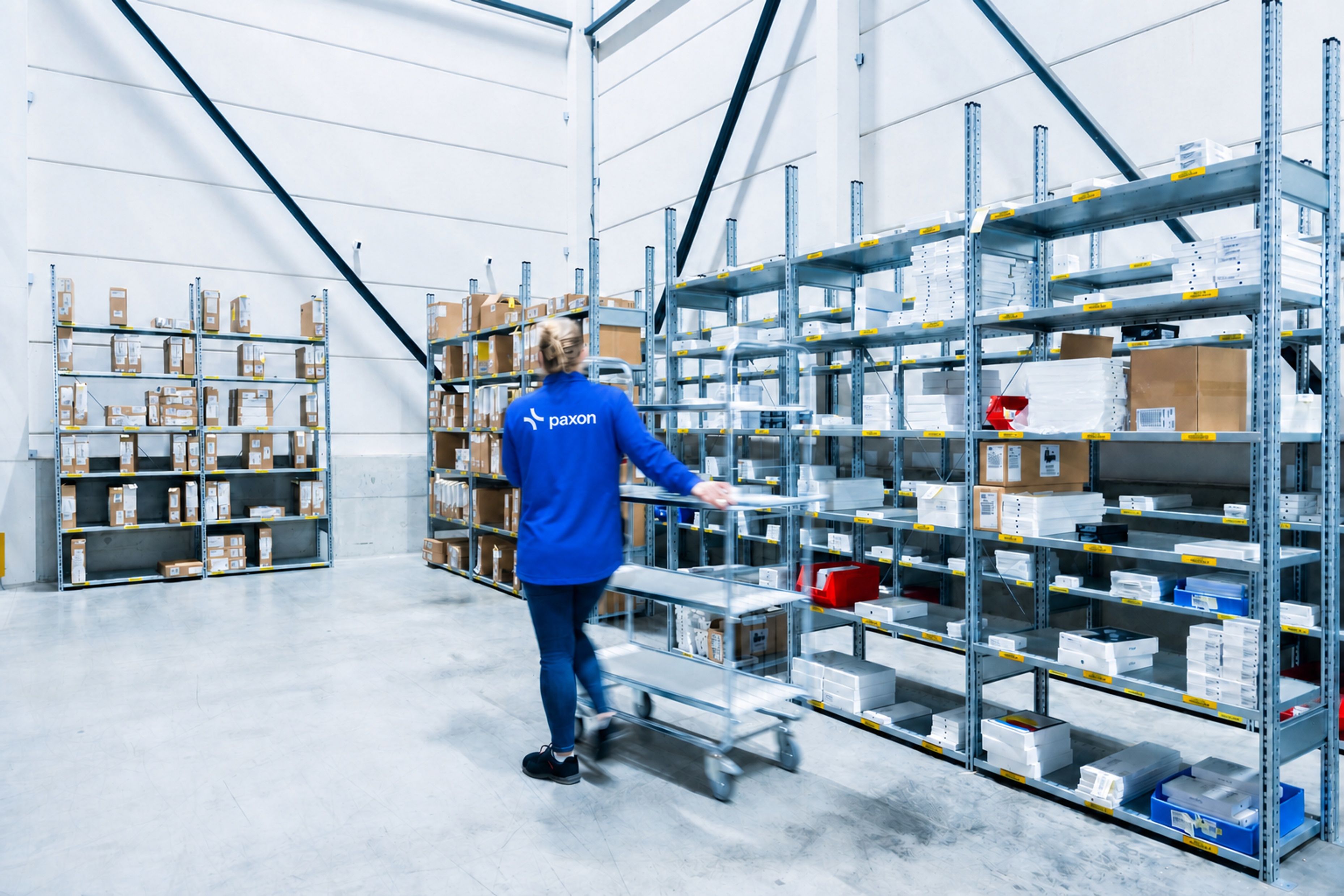 Une personne en chemise Paxon bleue pousse un chariot dans une allée d’entrepôt bordée d’étagères remplies de produits.