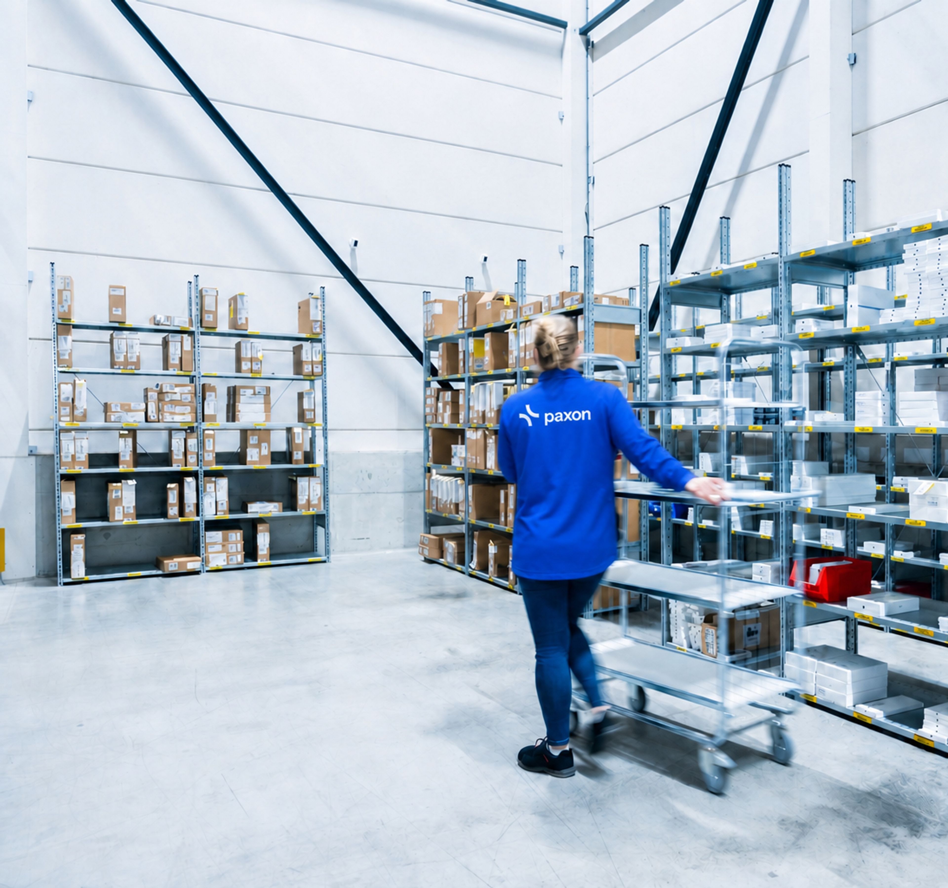 Une personne en chemise Paxon bleue pousse un chariot dans une allée d’entrepôt bordée d’étagères remplies de produits.
