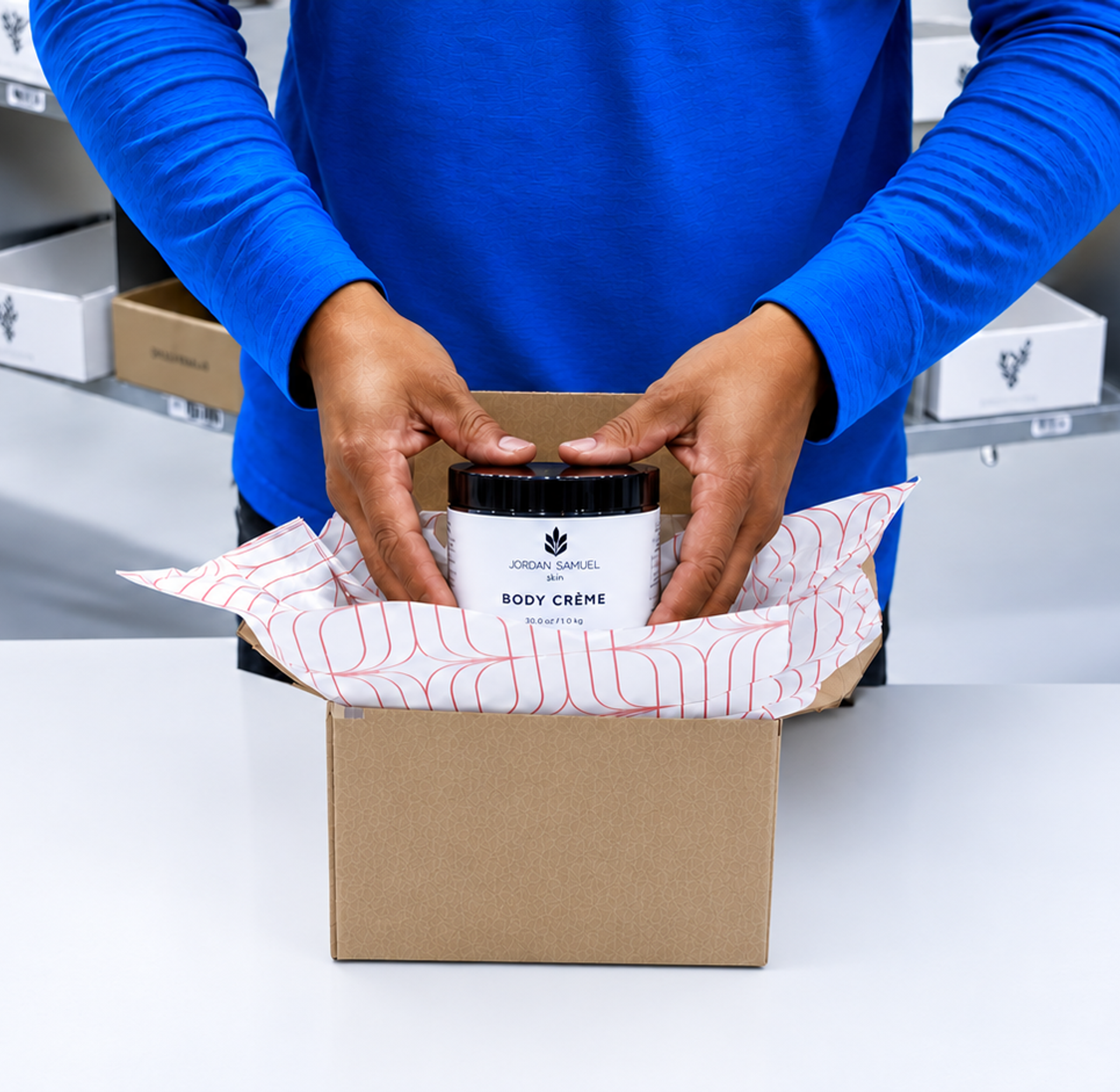 A person's hands place a jar of "Body Creme" into a small shipping box lined with tissue paper.