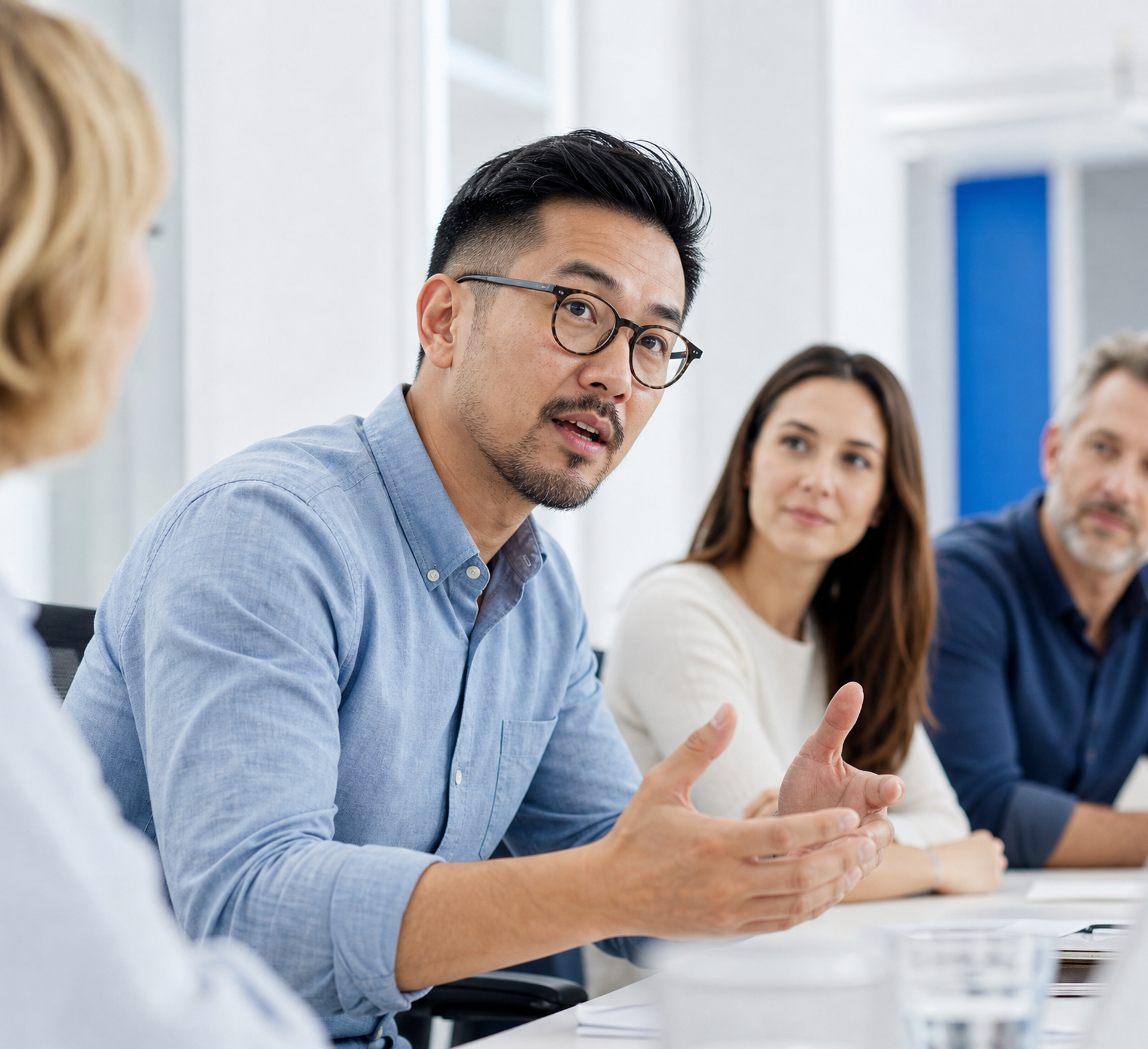 An Asian man speaks and gestures at a meeting table while others listen.