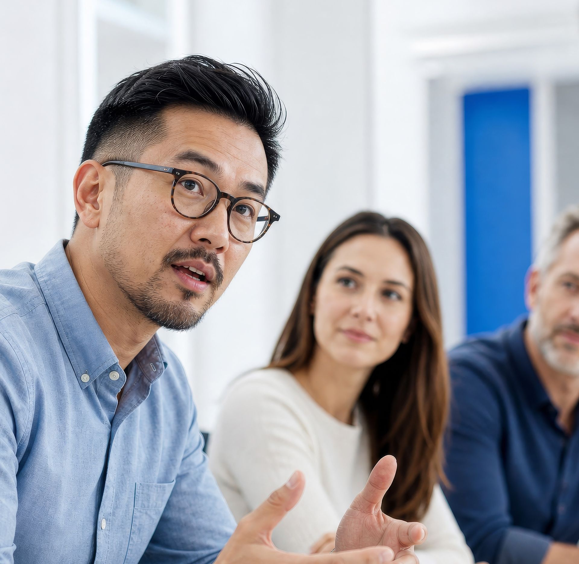 A man with glasses and a beard speaks and gestures during a meeting, with two colleagues listening.