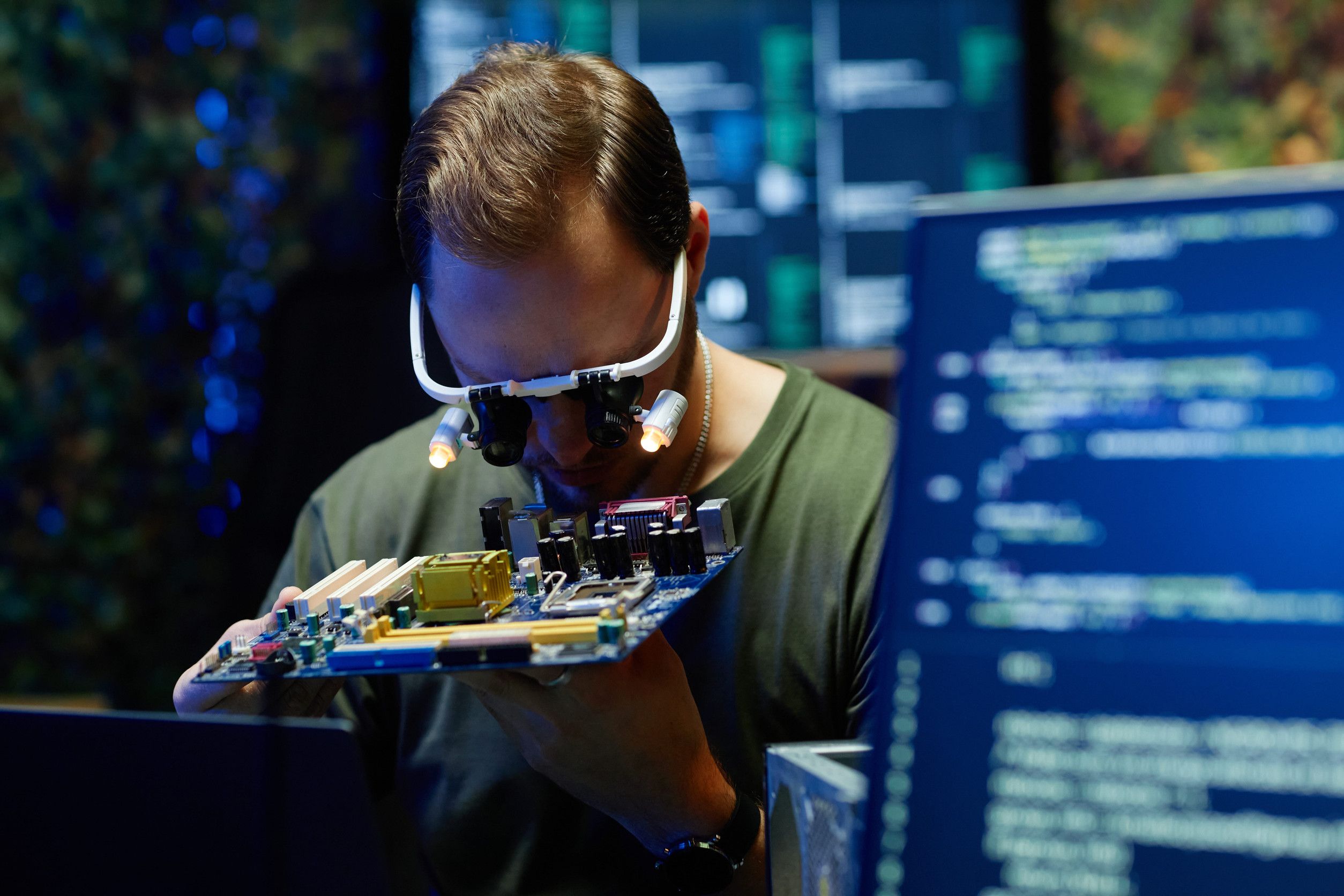 Un homme portant des loupes examine de près un circuit imprimé, avec un écran affichant du code à l’arrière-plan.
