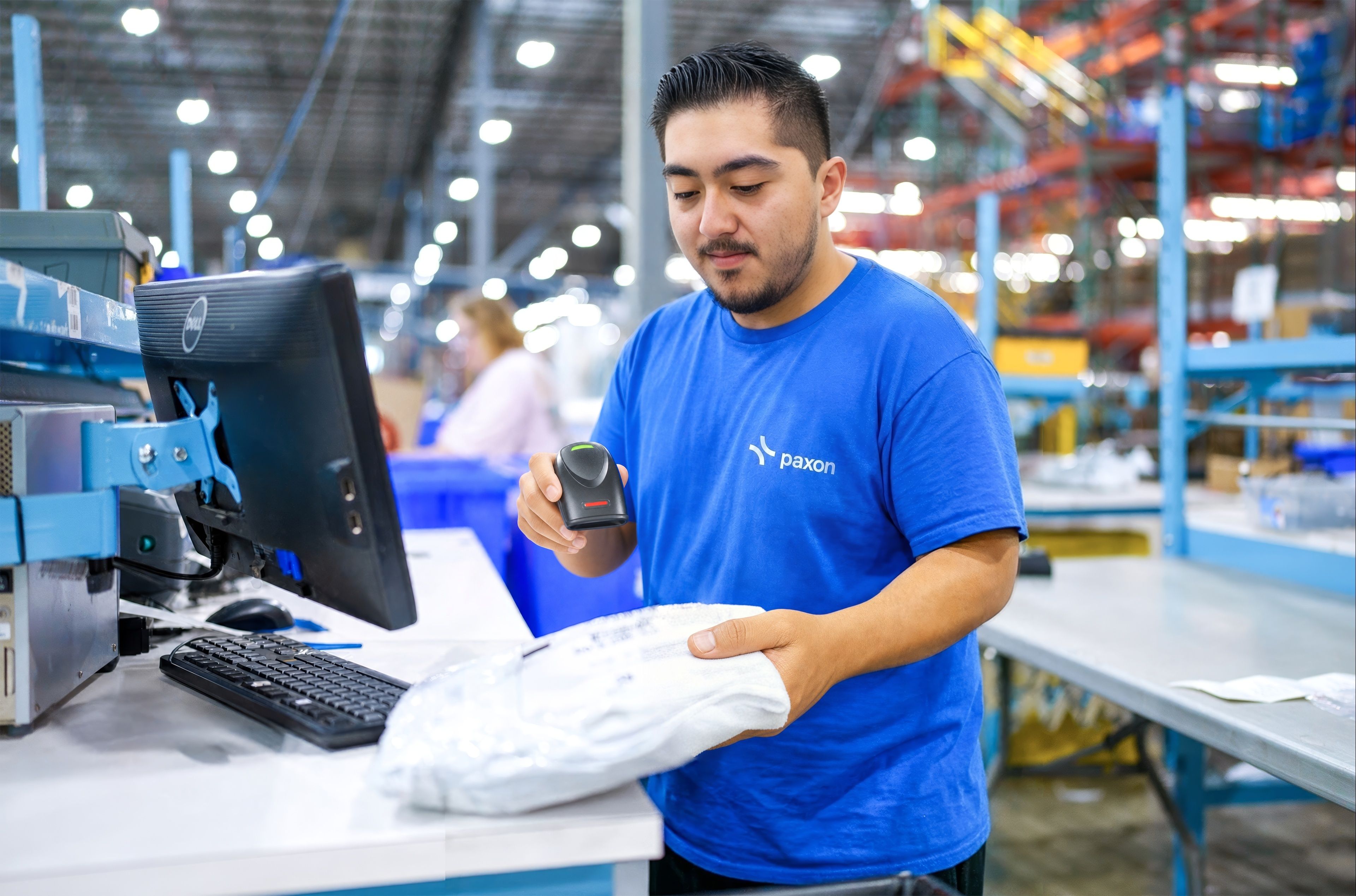 A man in a blue "paxon" t-shirt scans a white package at a workstation in a warehouse.