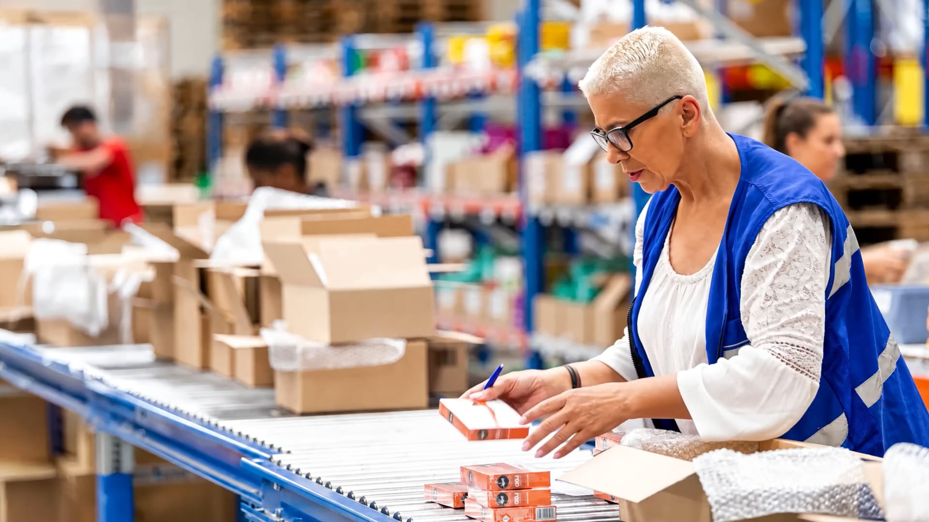 Woman in a blue vest checking packages on a warehouse conveyor belt.