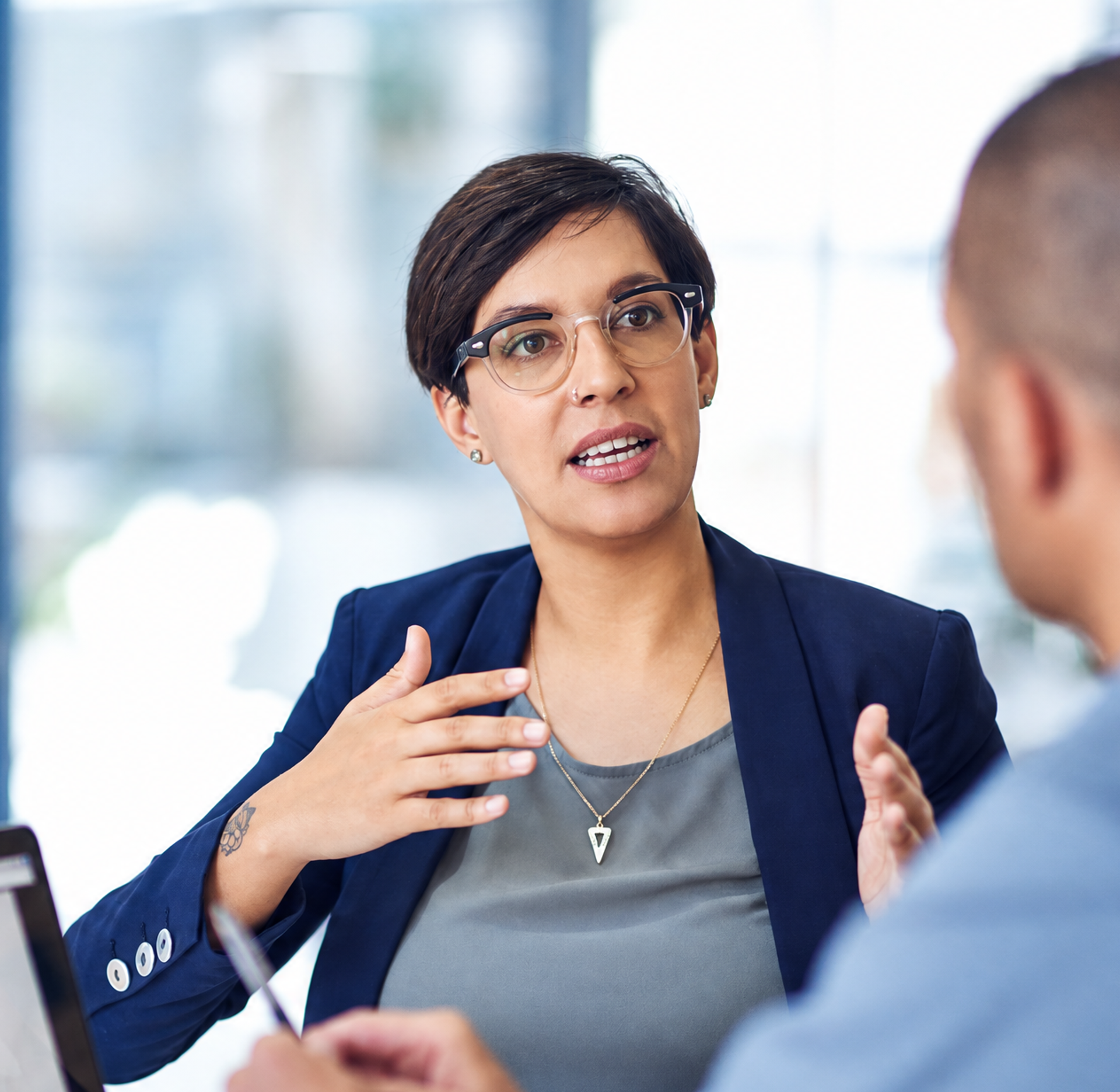 A woman in a blazer and glasses gestures while speaking to a colleague in an office meeting.