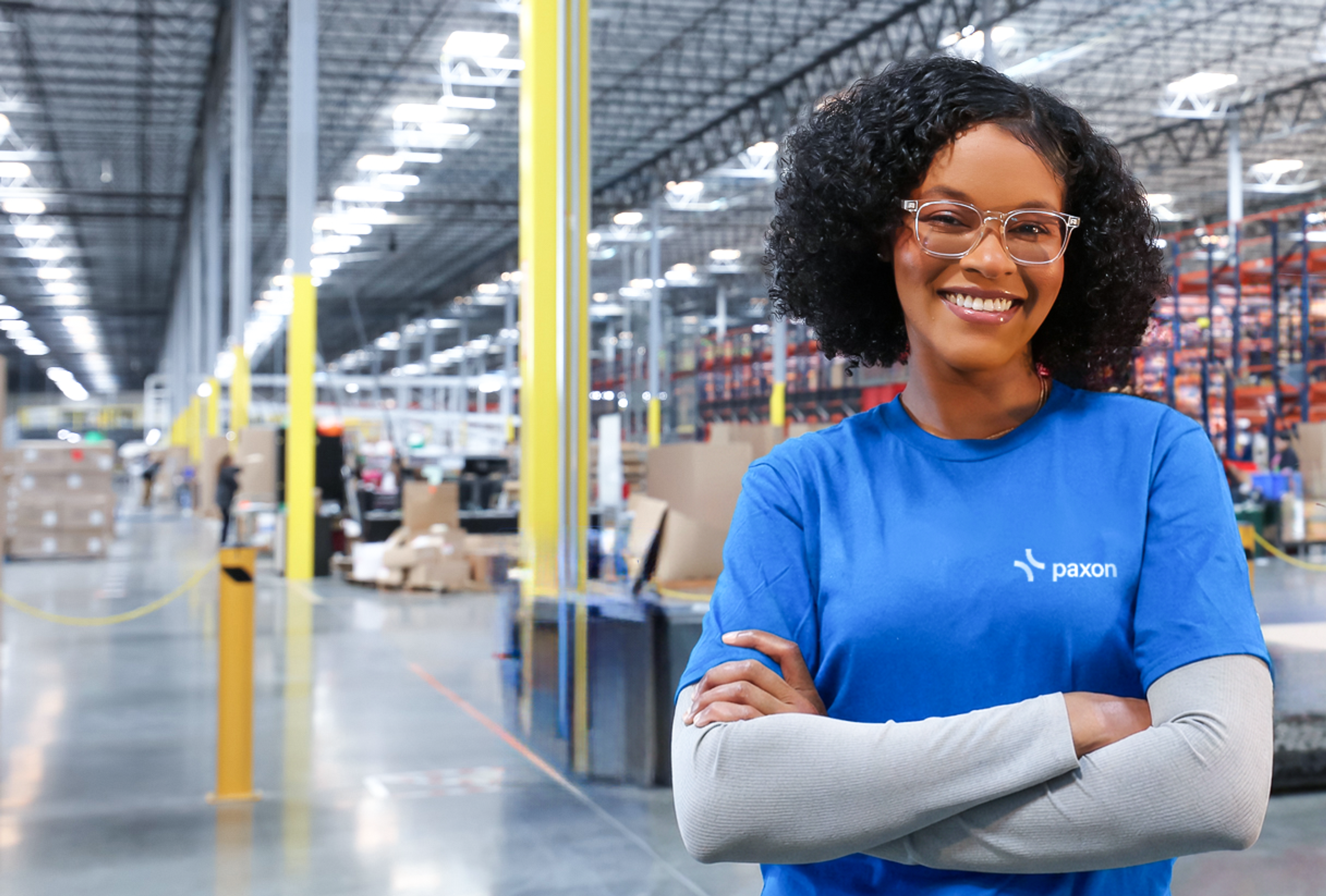 Smiling Black woman in a blue Plexon t-shirt with crossed arms in a large warehouse.